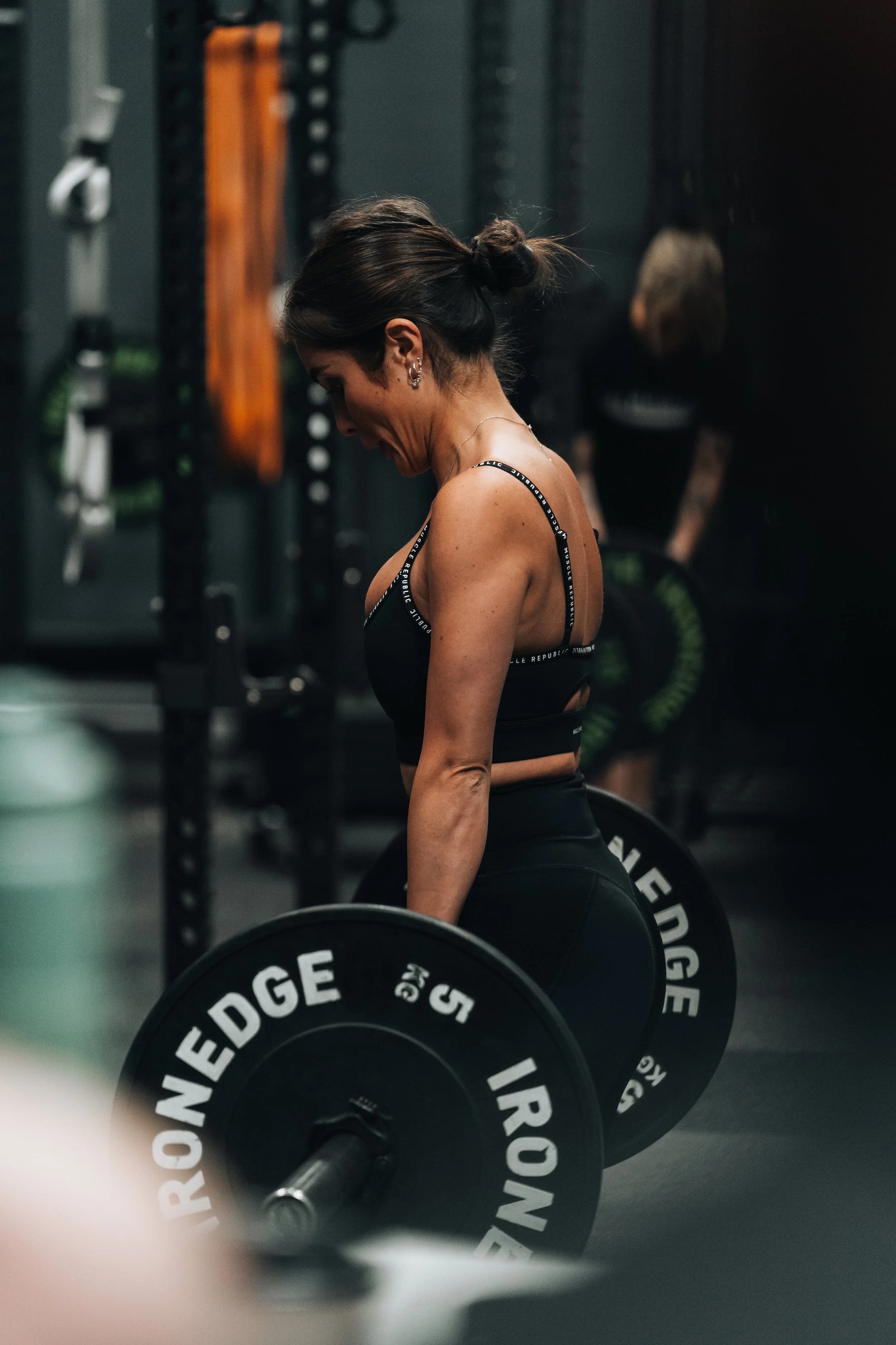 A woman with brown hair tied in a bun, wearing a black sport bra, is at the gym lifting a barbell with weight plates labeled EDDGE. She is in front of black wall-mounted gym equipment with various workout tools.