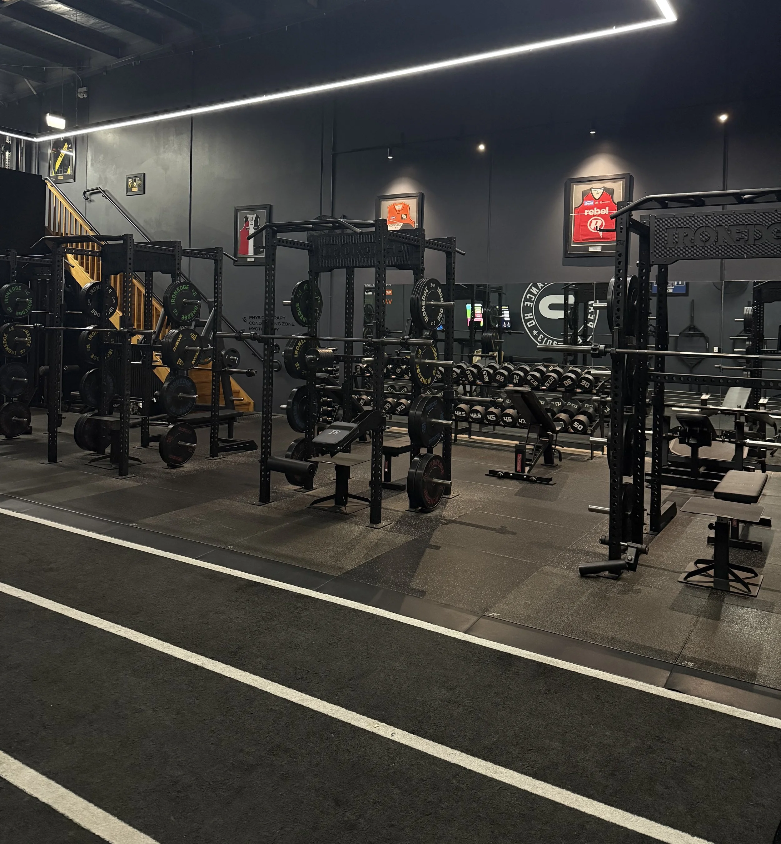 Interior of a gym with workout equipment including weight racks, benches, and framed jerseys on the walls, illuminated by overhead lighting.