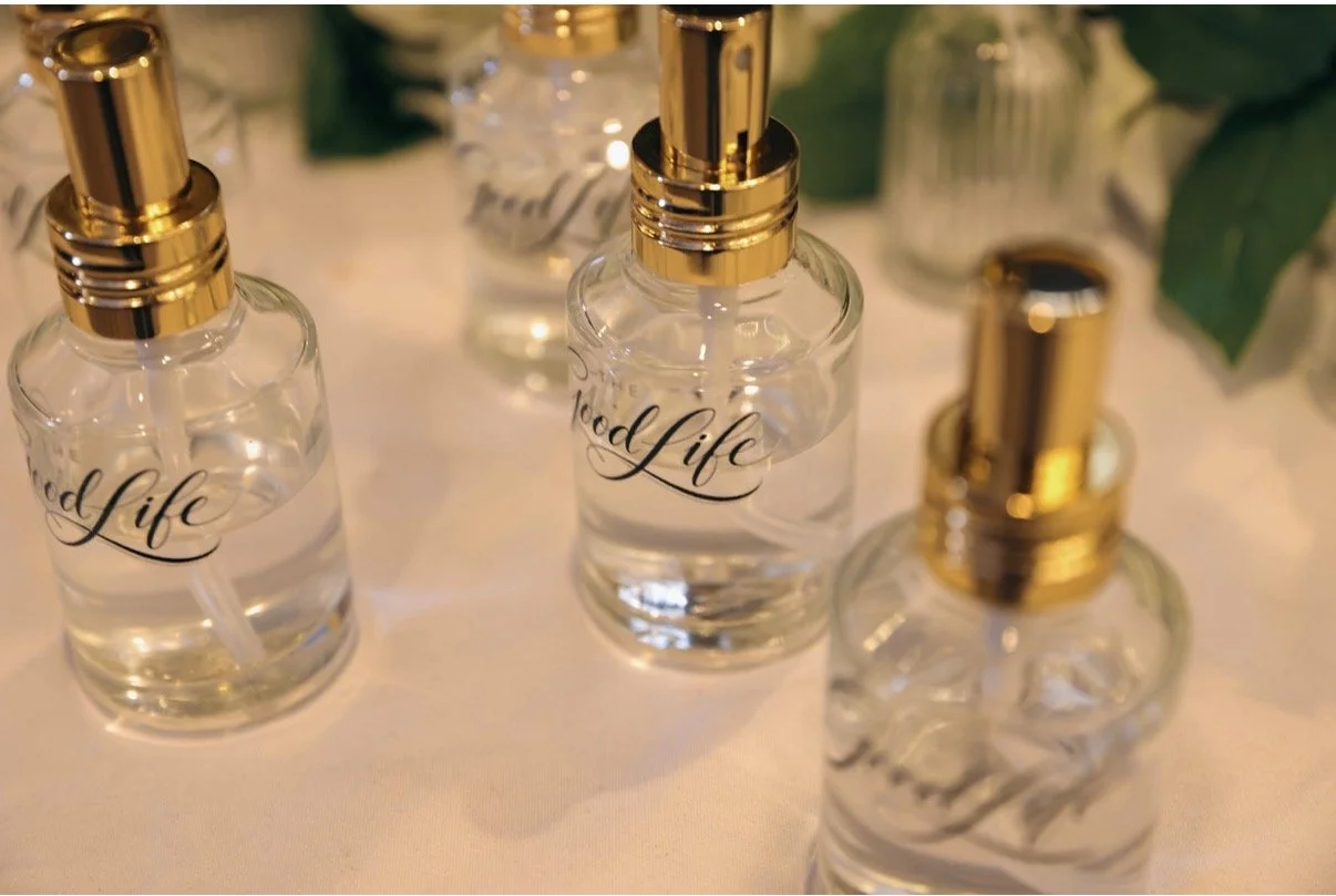 Several clear glass spray bottles with black and gold caps and sprayers, labeled 'Good Life,' arranged on a white surface with green leaves and additional bottles in the background.