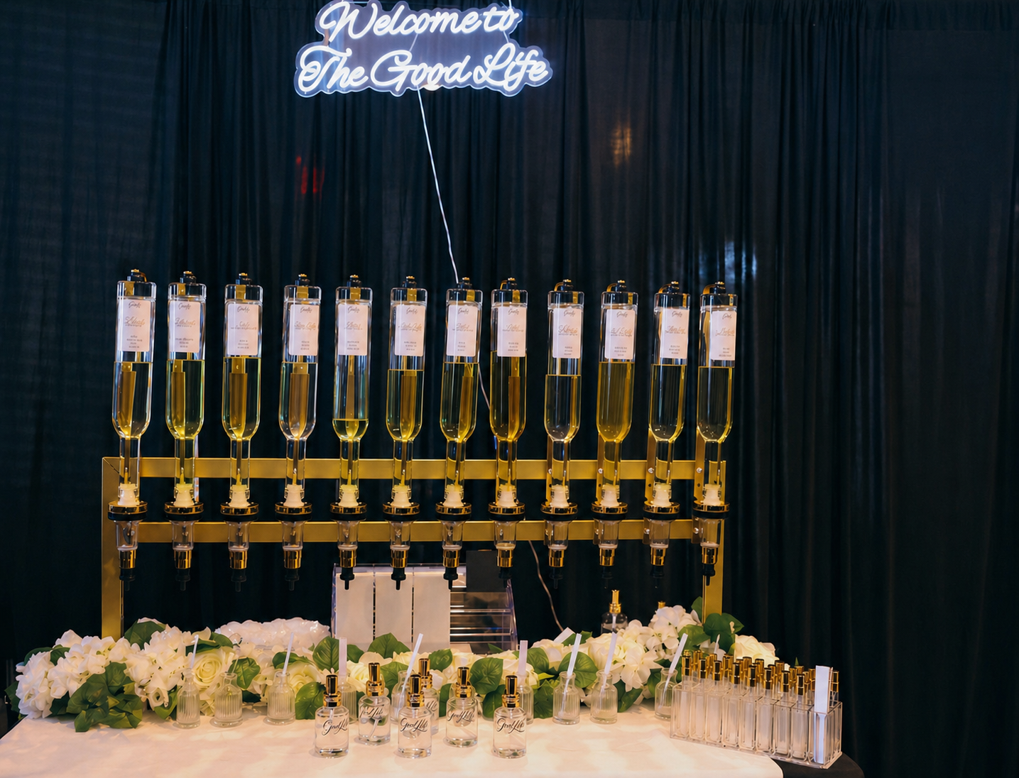 A decorative display of perfume bottles on a table with white flowers and greenery, featuring a neon sign that reads 'Welcome to The Good Life' in the background.