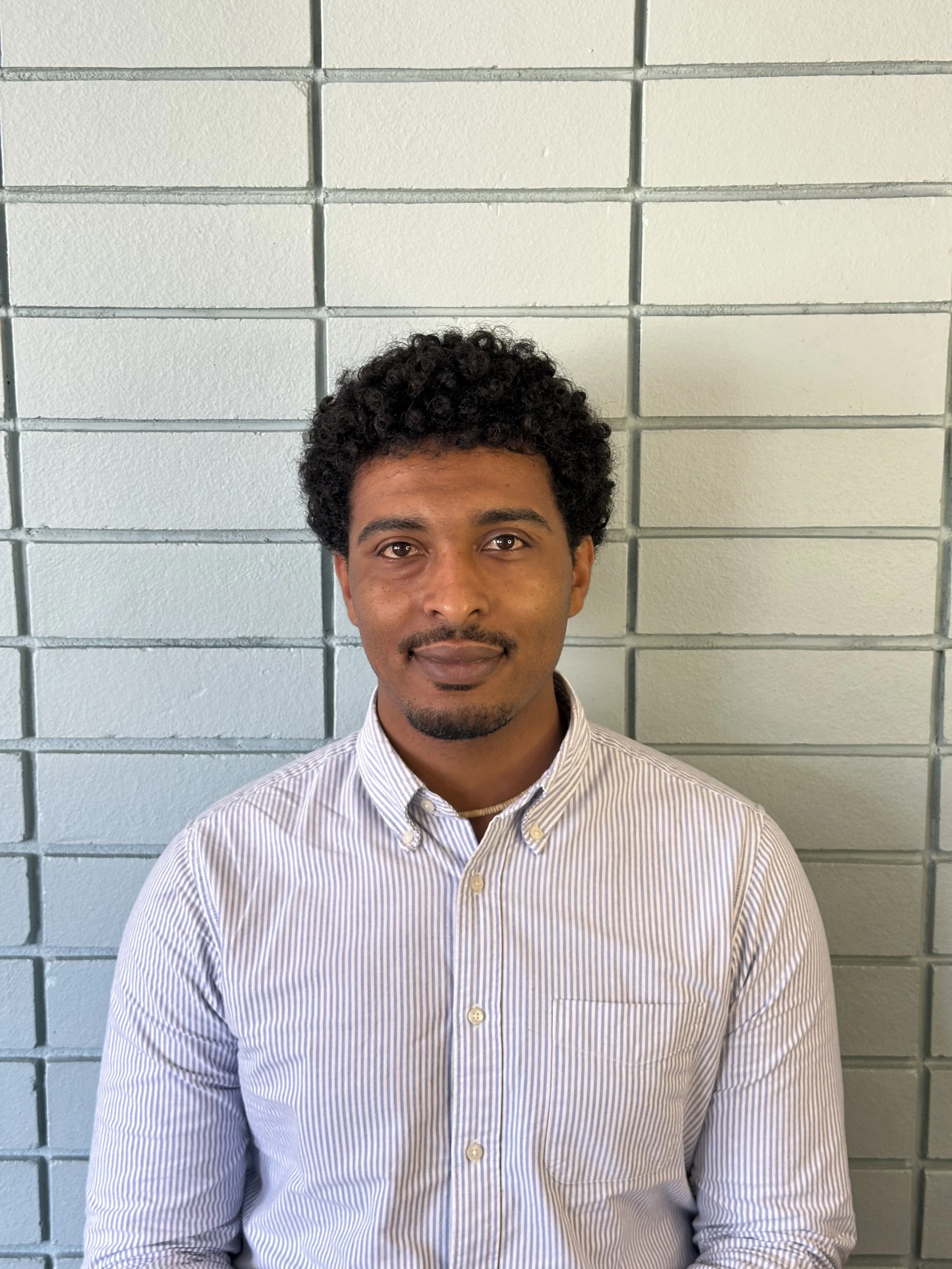 Portrait of a smiling man with curly hair and a beard, wearing a collared striped shirt, standing in front of a brick wall.