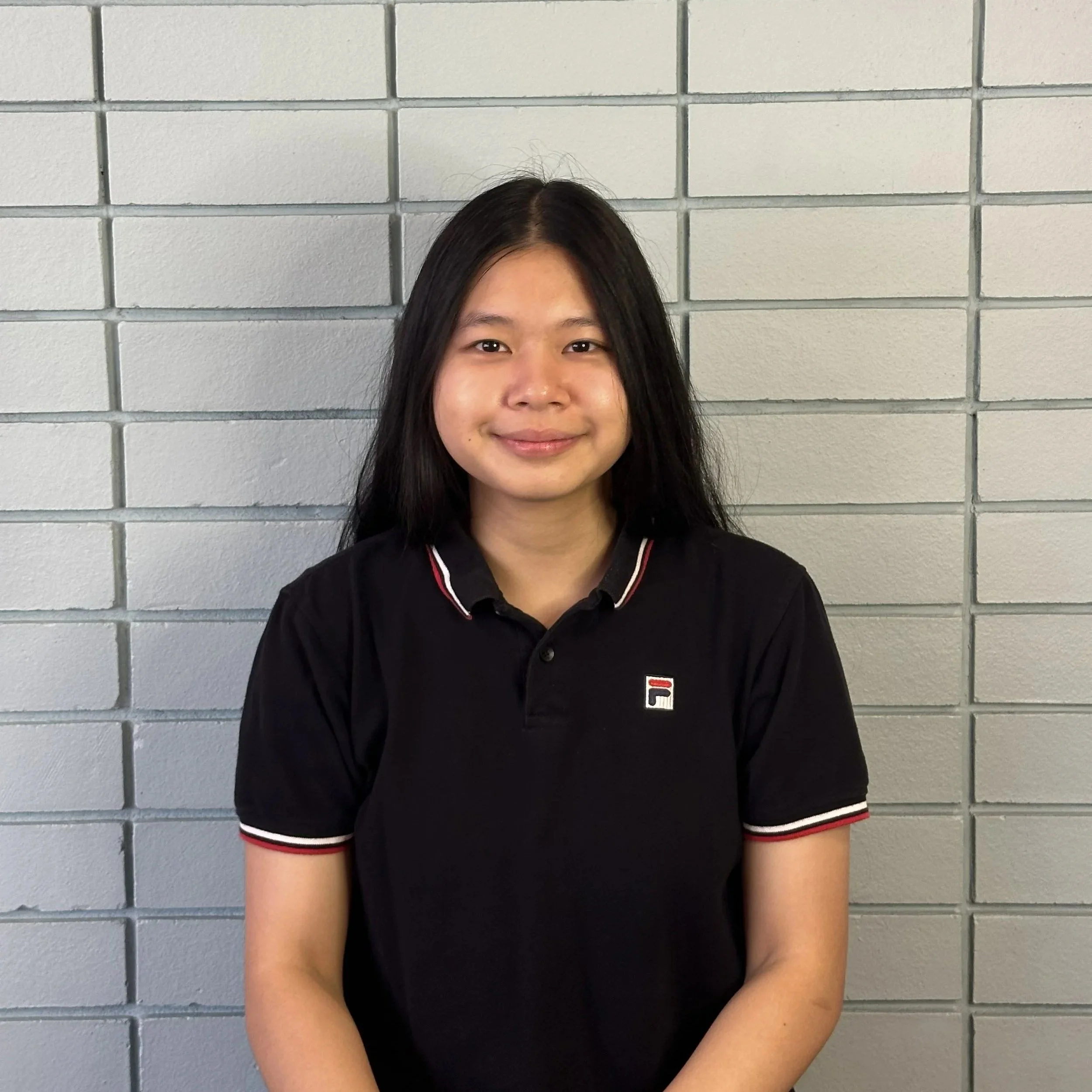 A woman with black hair, wearing a collared shirt, sitting against a brick wall background. 