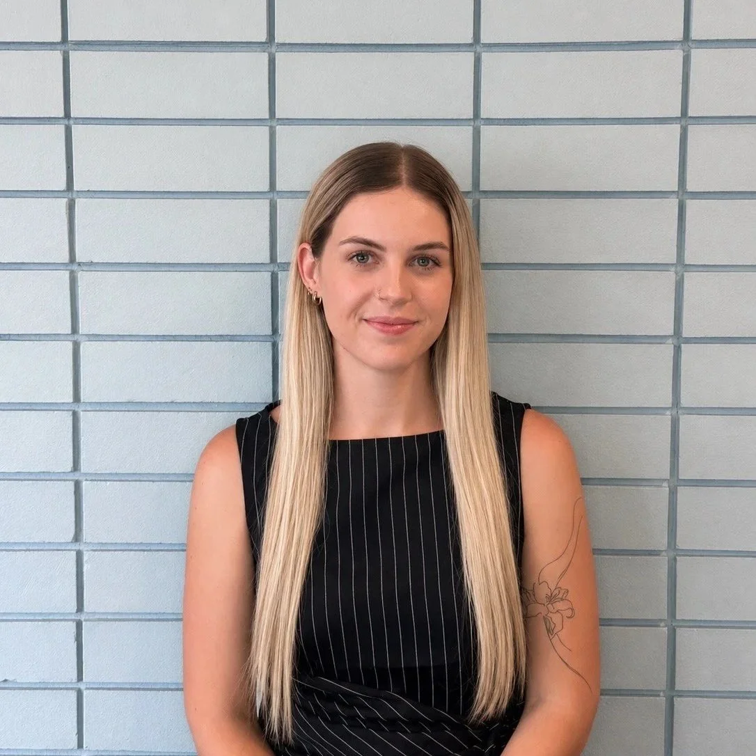 A woman with long blonde hair, wearing a striped shirt, sitting against a brick wall background.