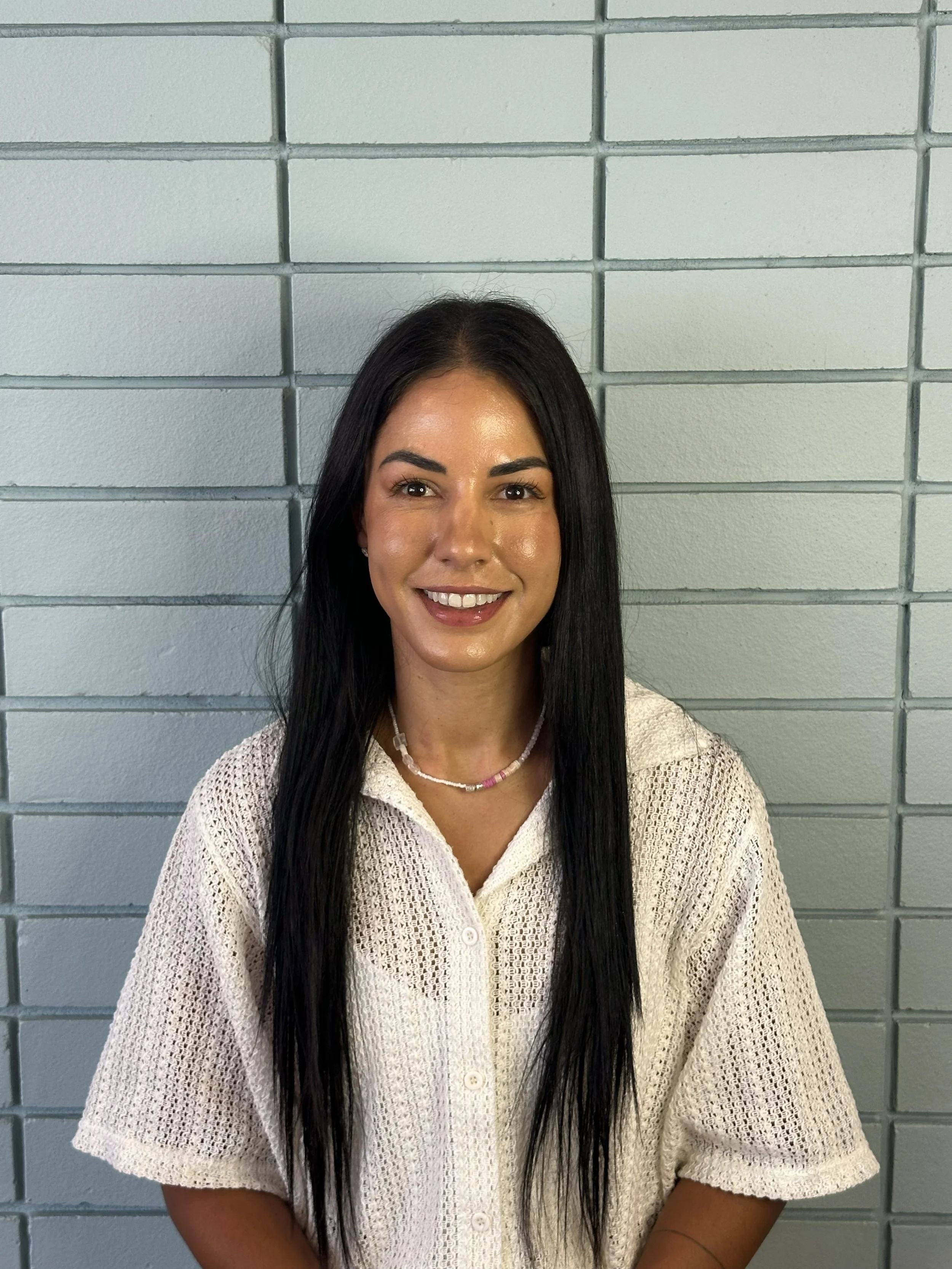 A woman with long dark hair, smiling, dressed in a sweater, standing in front of a brick wall.