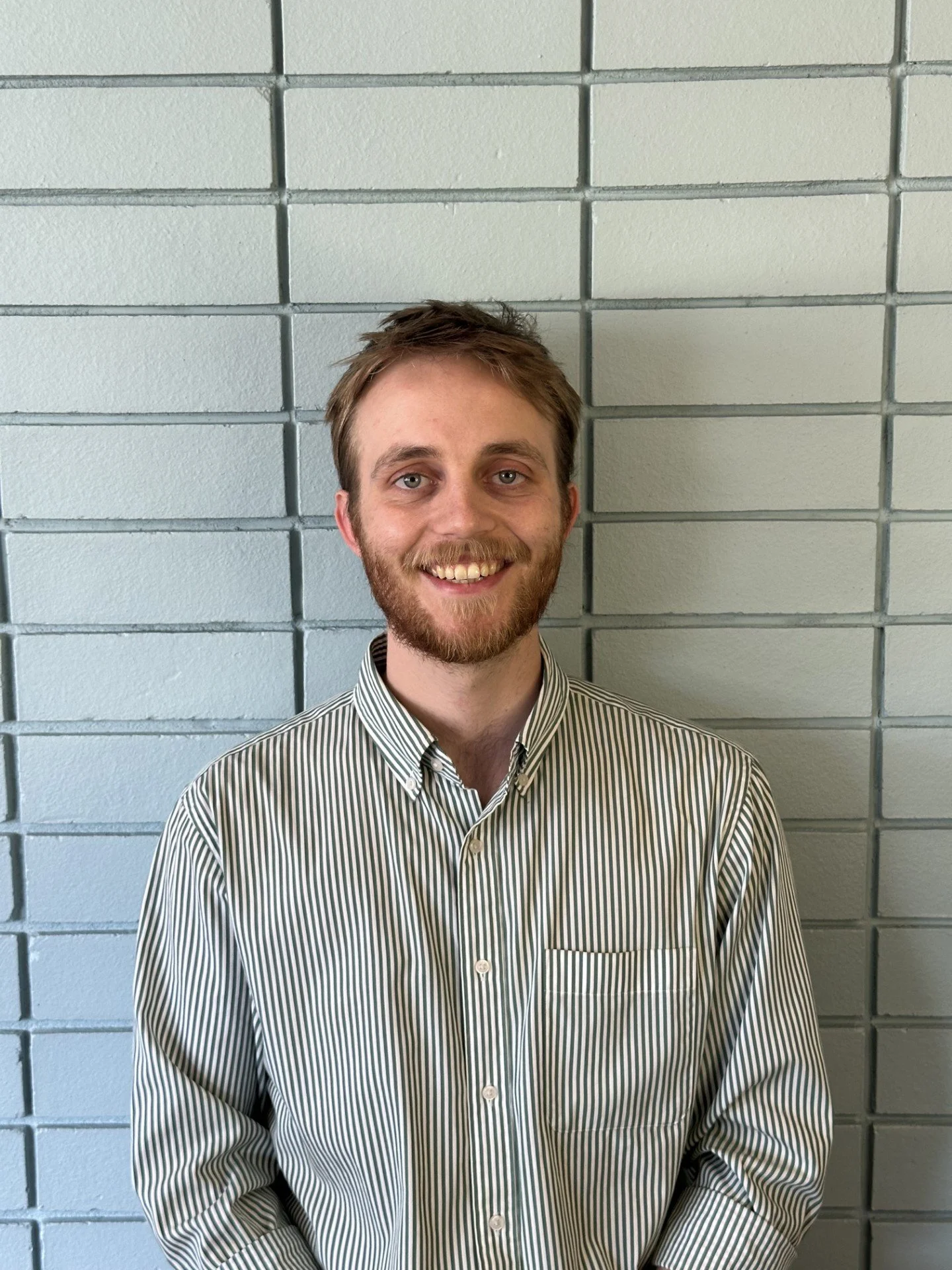 Portrait of a young man with short hair, mustache, smiling, standing in front of a brick wall.