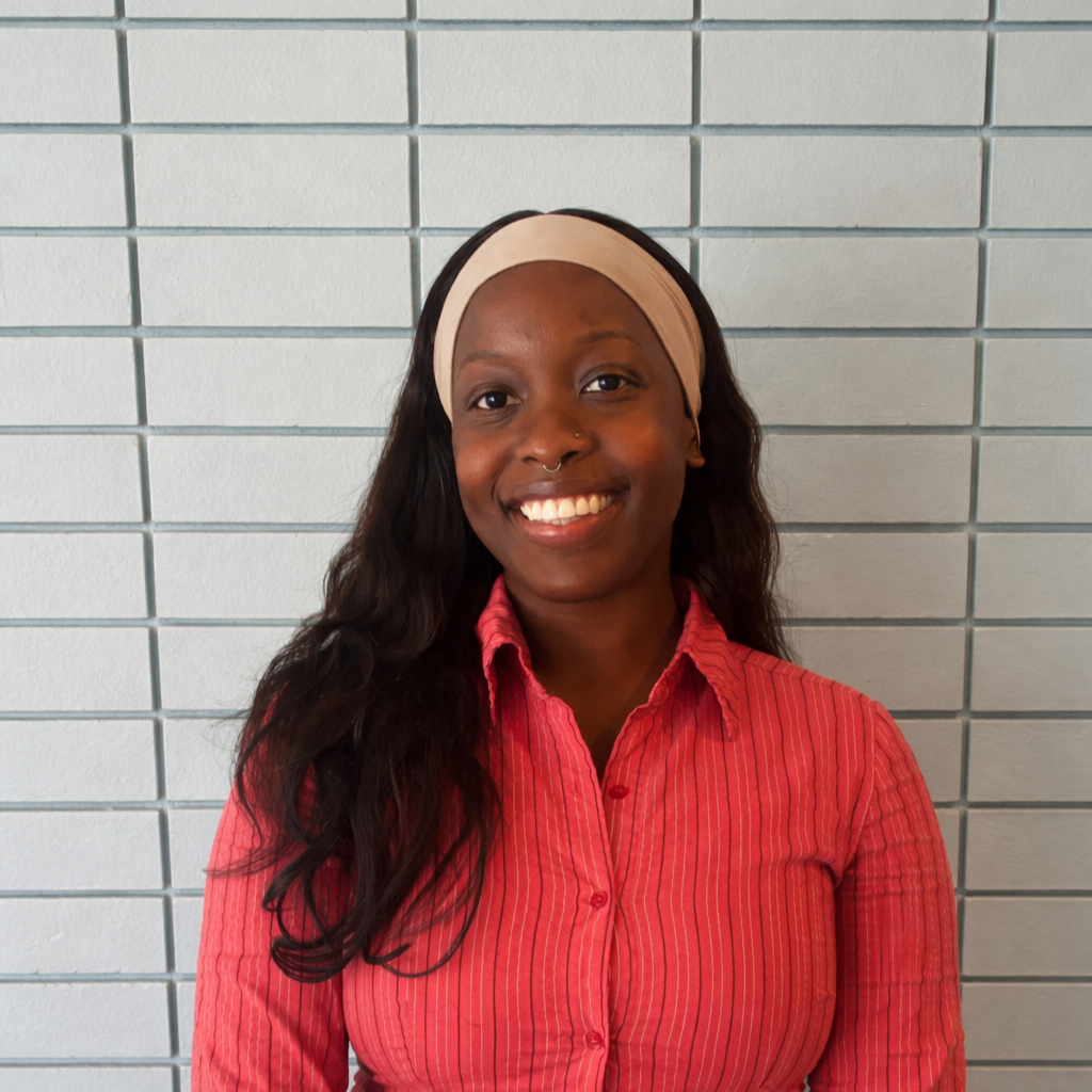 A woman with black hair, wearing a headband and striped shirt, sitting against a brick wall background. 