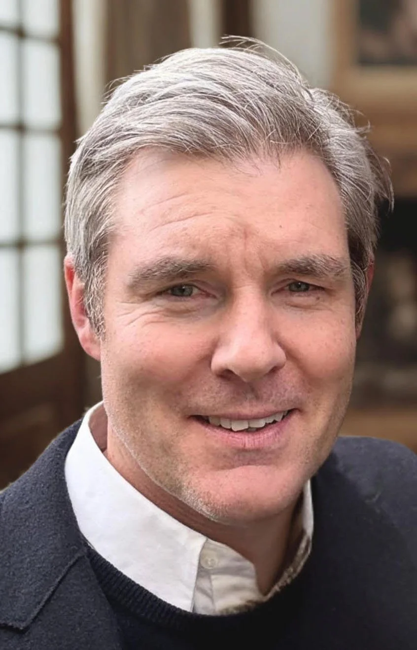 Close-up of a smiling middle-aged man with gray hair, wearing a white shirt and dark sweater, indoors with a window in the background.