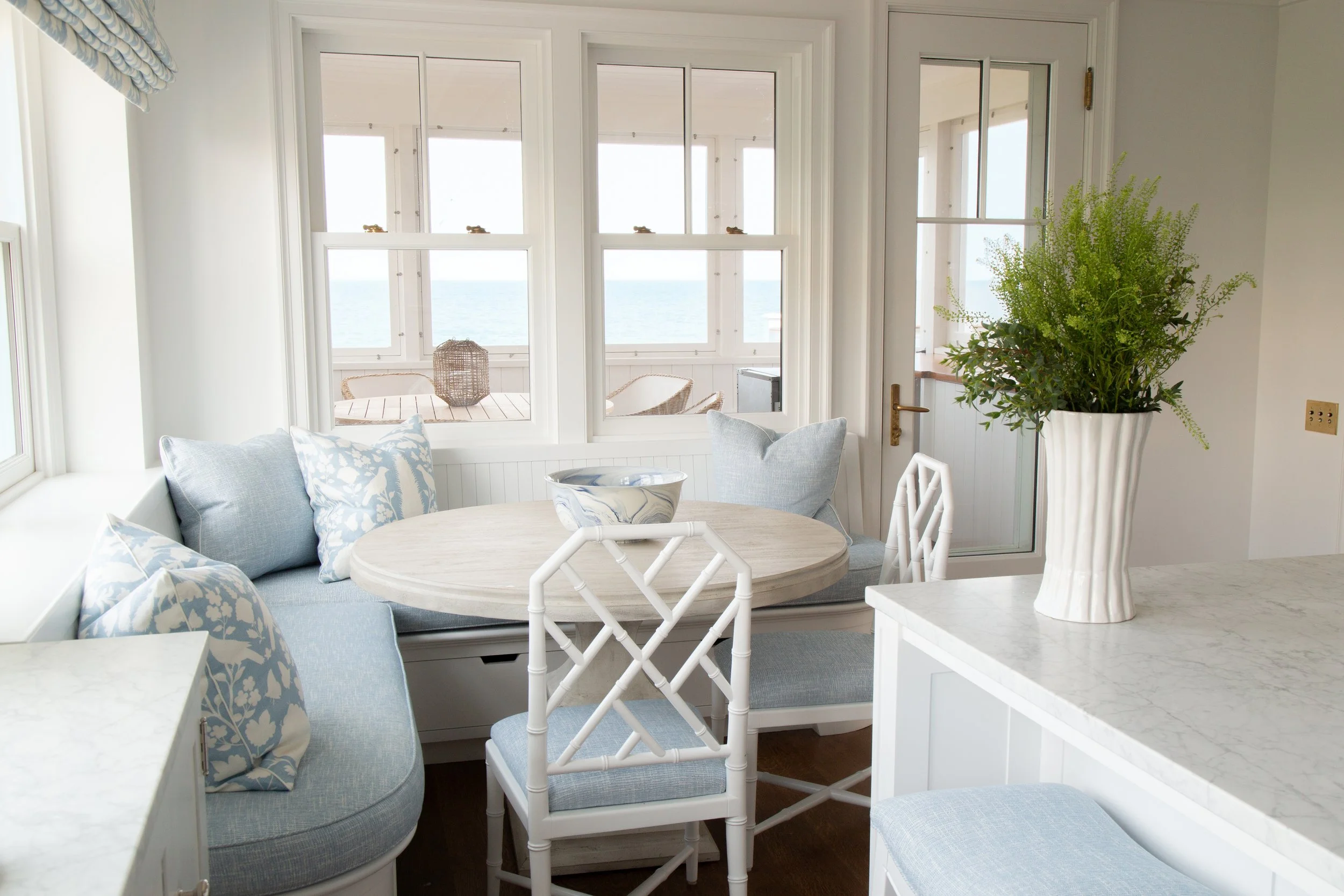 Cozy dining nook with a round table, cushioned chairs, decorative pillows, and large windows overlooking the ocean, with a potted plant on the counter.