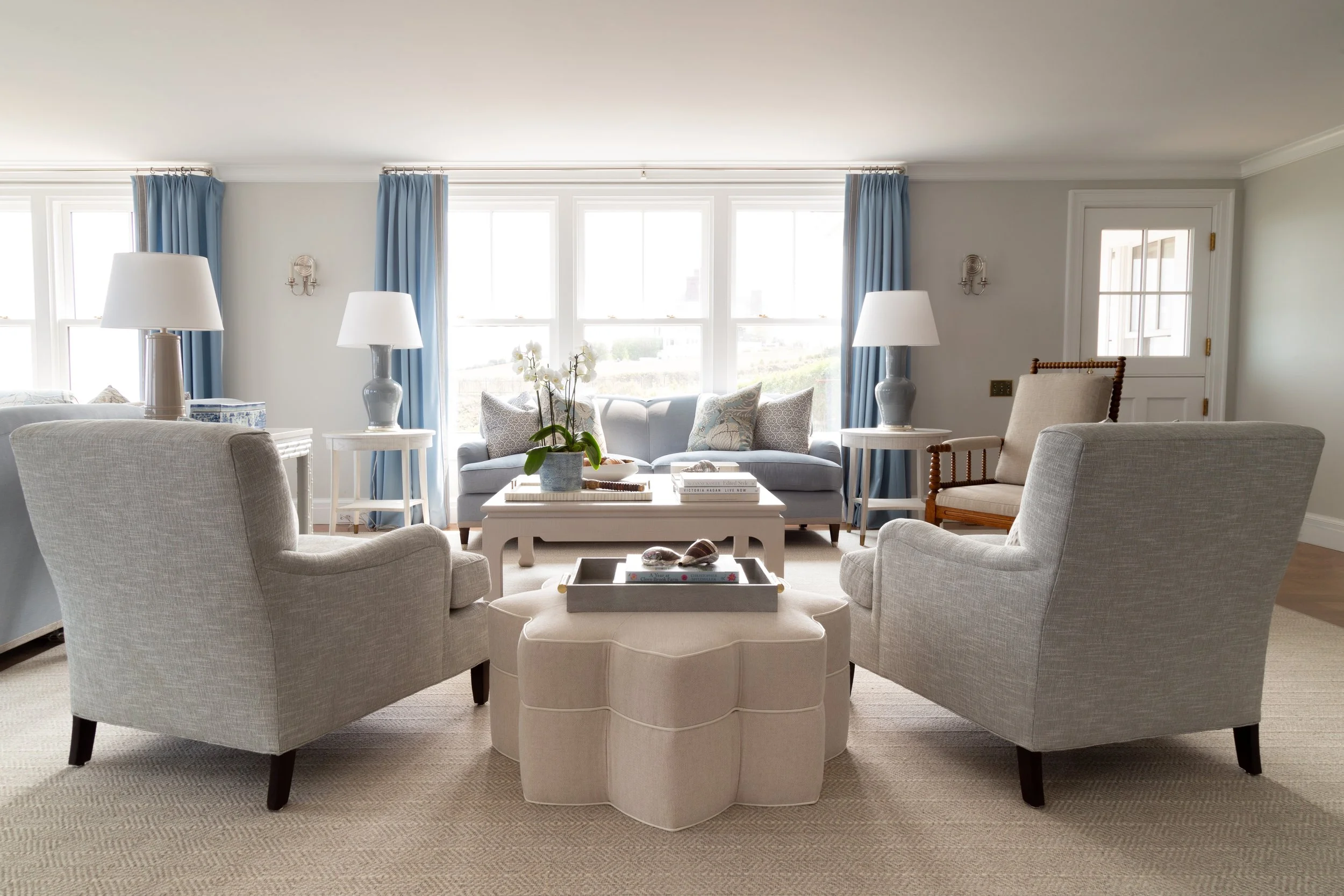 Living room with two gray armchairs around an ottoman, a sofa, two lamps, and large windows with blue curtains.