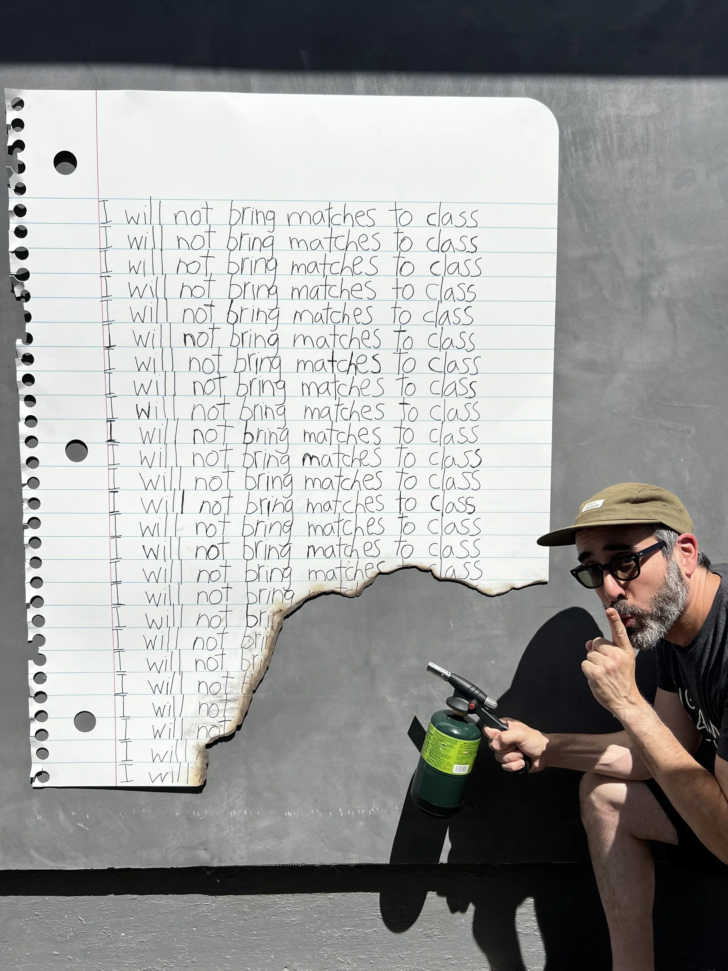 A large torn-out sheet of notebook paper with the repeated phrase 'I will not bring matches to class' written multiple times. A man with glasses and a beard, wearing a baseball cap, is crouched next to the paper, holding a small torch or lighter and making a shushing gesture.