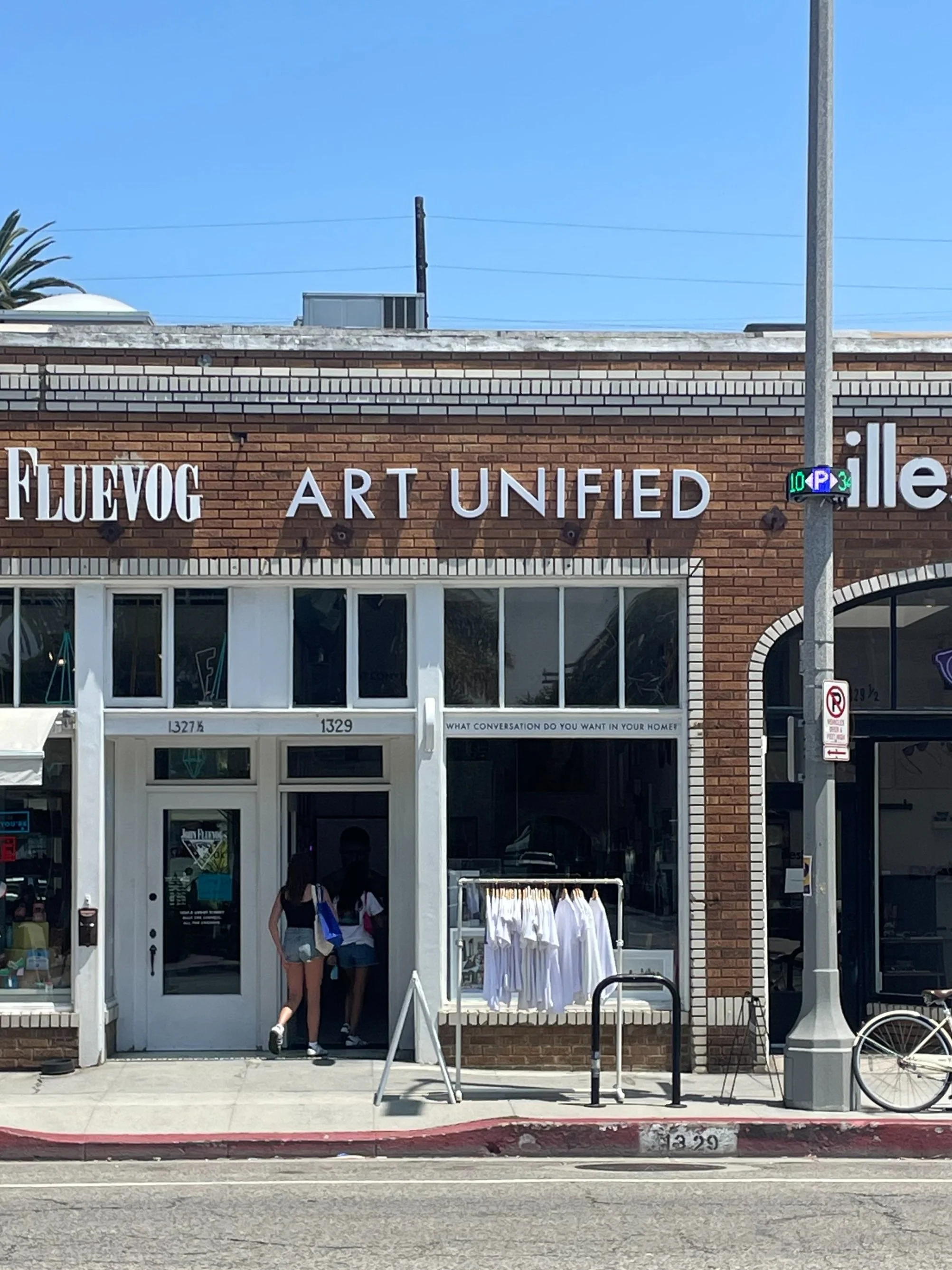 storefront with signs reading 'FLUEVOG' and 'ART UNIFIED' on a brick building, with two people entering and white clothing displayed outside.