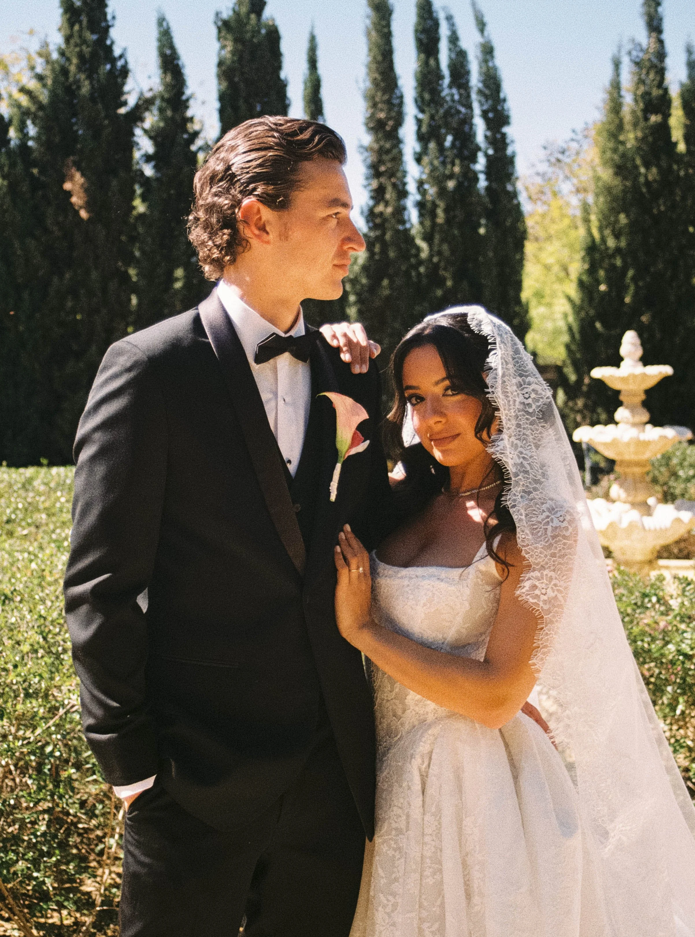 Editorial Italy wedding photographers bride and groom standing close together outdoors, with trees and a fountain in the background. The groom is wearing a black tuxedo with a bow tie, and the bride is wearing a white lace wedding dress with a veil.