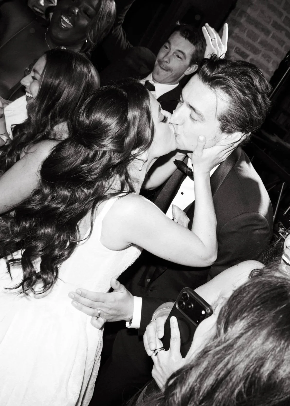 A black and white photo of a couple kissing, surrounded by friends at a celebration or party, with one woman holding a phone.