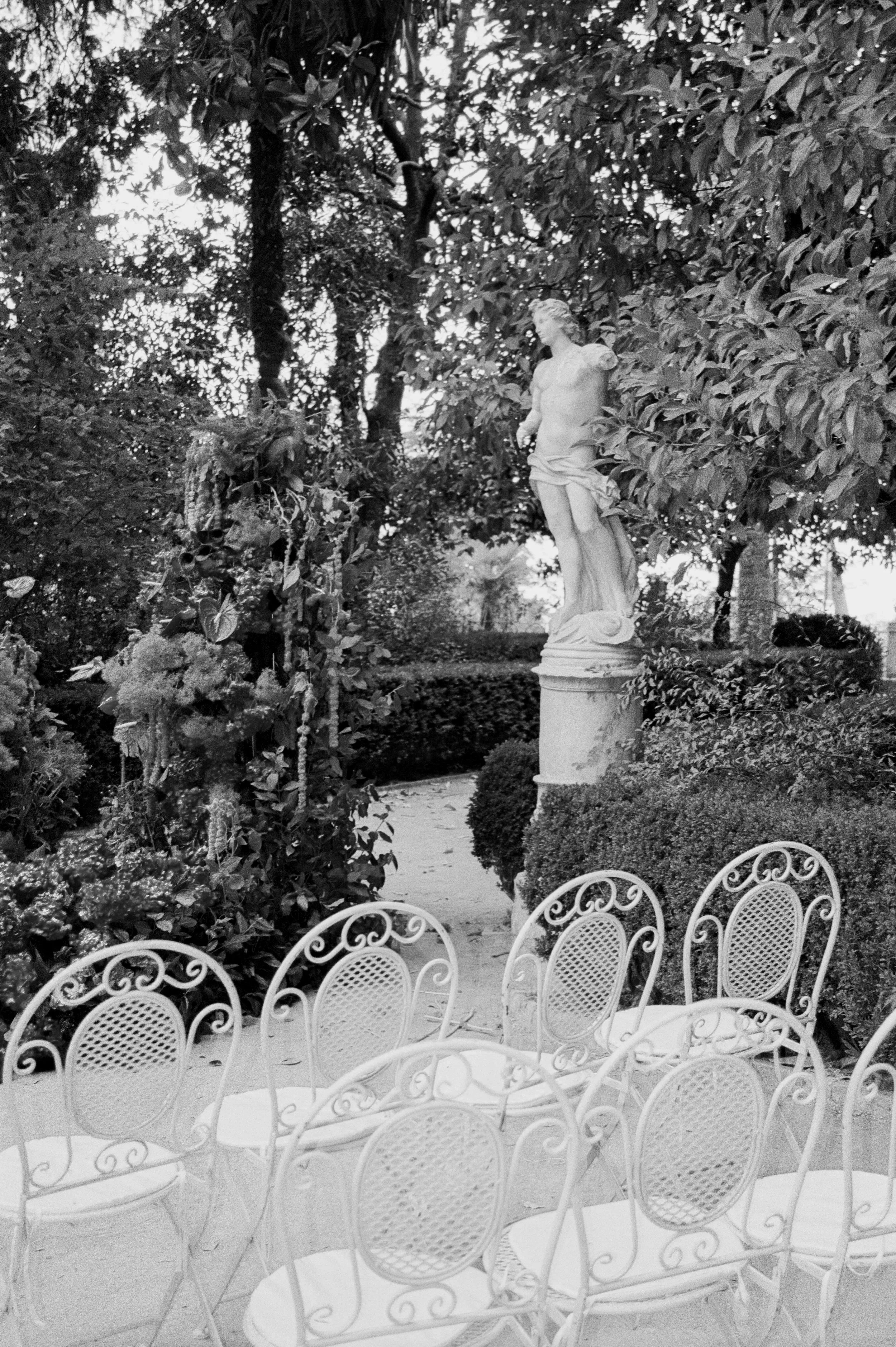 Garden scene with a white ornate metal table and chairs, a classical sculpture of a male figure, surrounded by lush trees and plants.