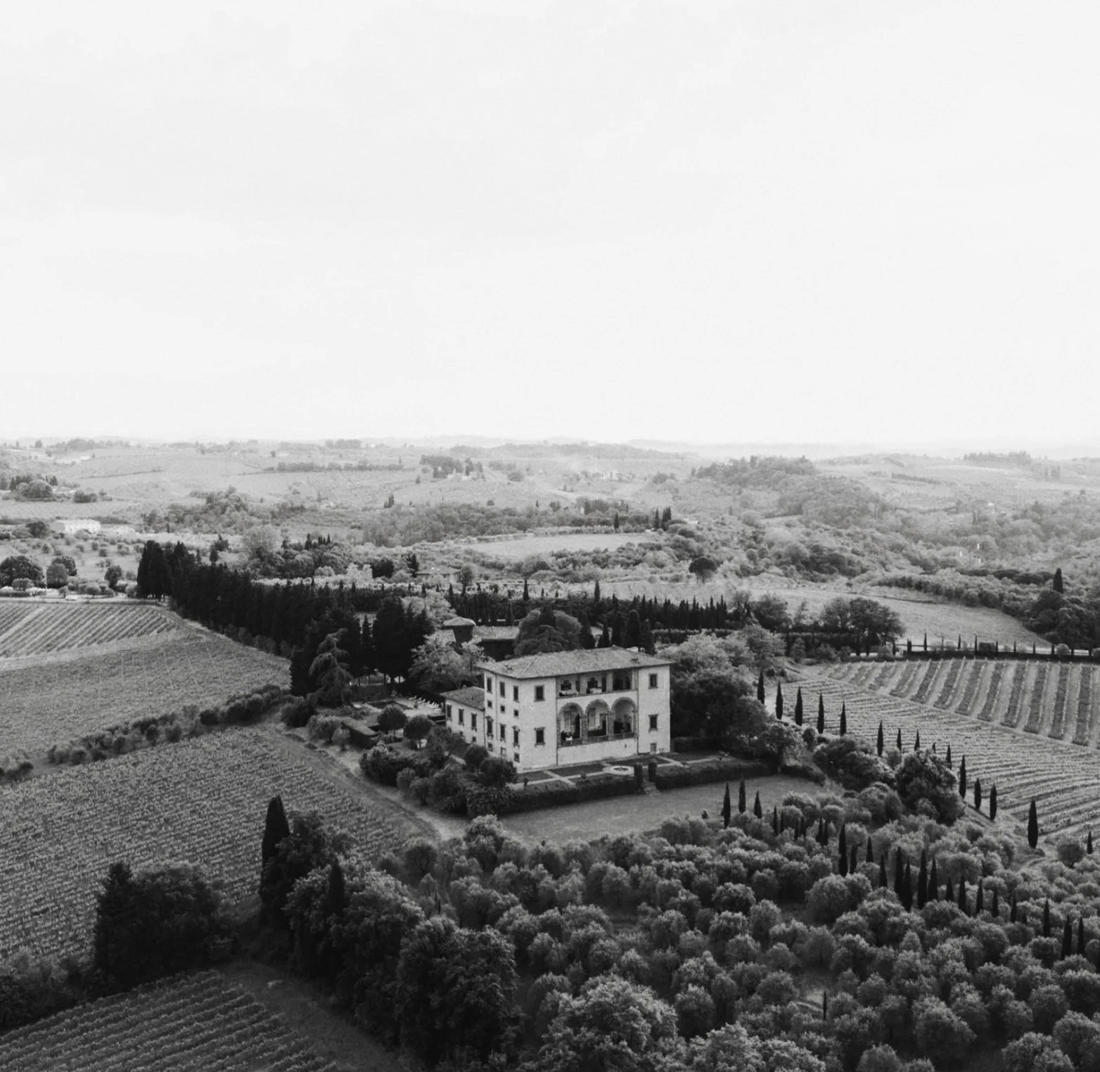 A black and white landscape view of rolling hills with fields, trees, and a white Mediterranean-style villa in the foreground by Italy wedding photographer with drone
