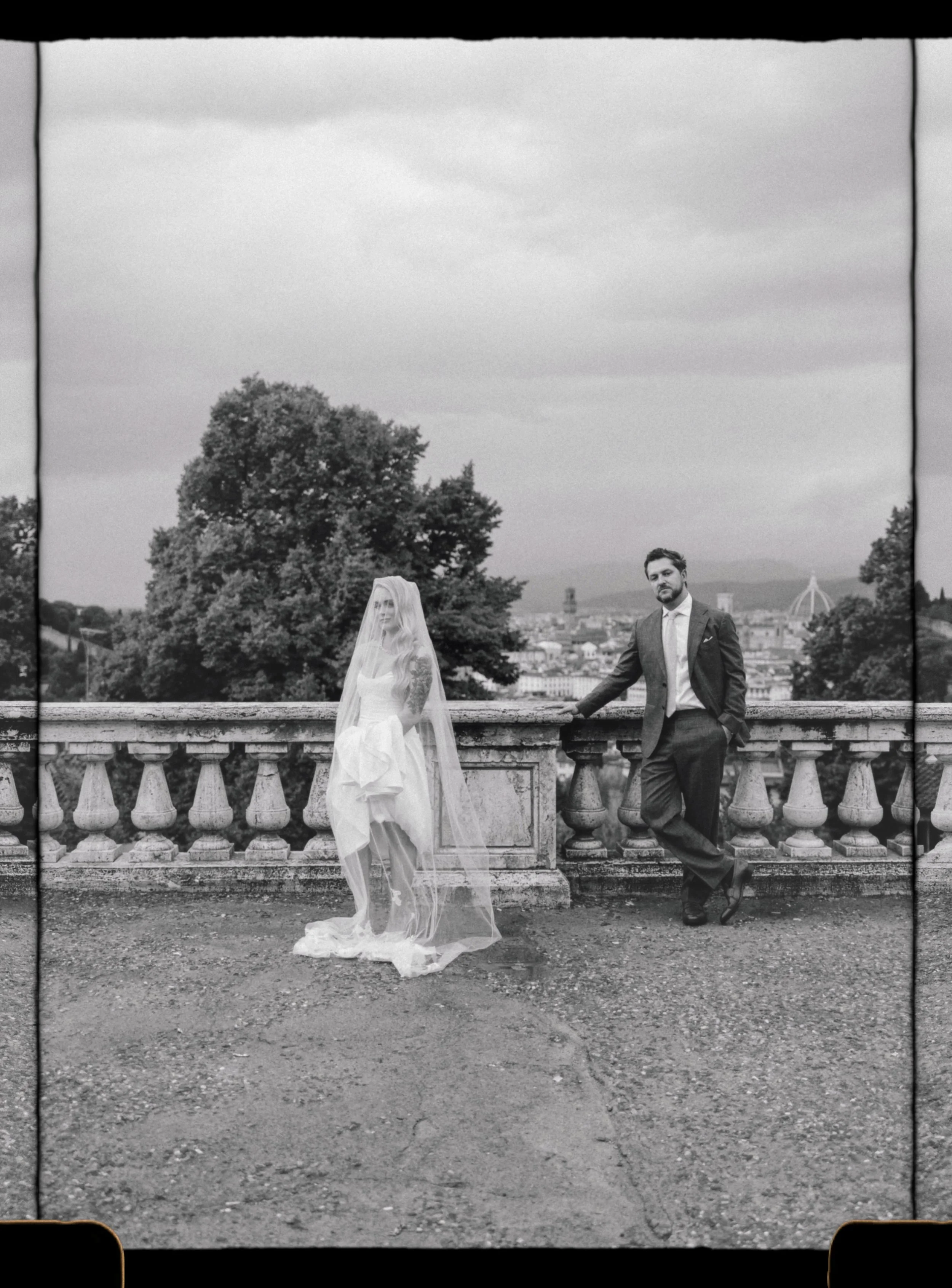 A black and white photo of a bride and groom standing on a stone balcony with trees and cityscape in the background captured by editorial wedding photographer in Tuscany in Piazzale Michelangelo