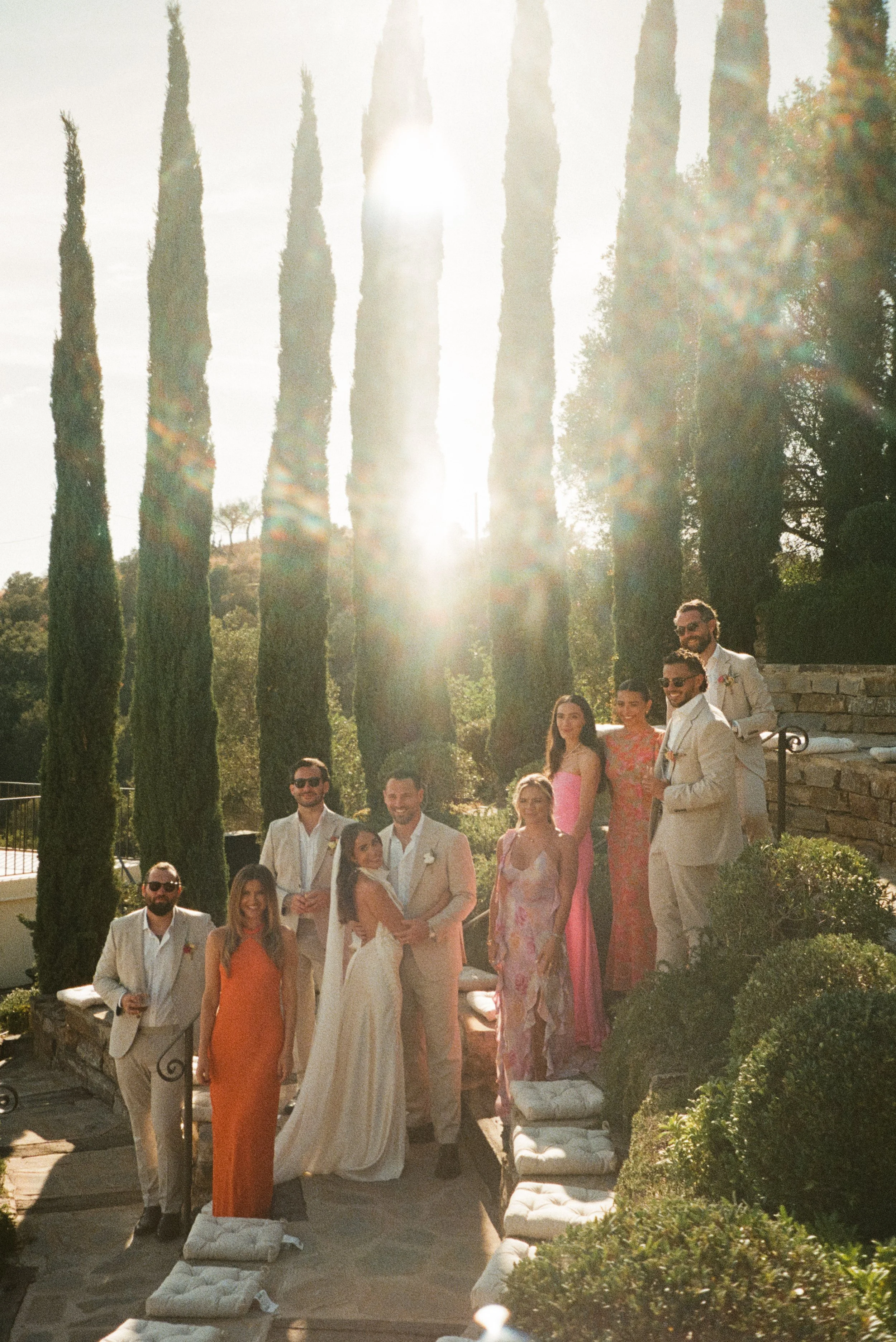 Italy wedding photographer captures group of bridal party dressed in formal and colorful attire standing outdoors on a sunny day, with tall trees and steps in the background.