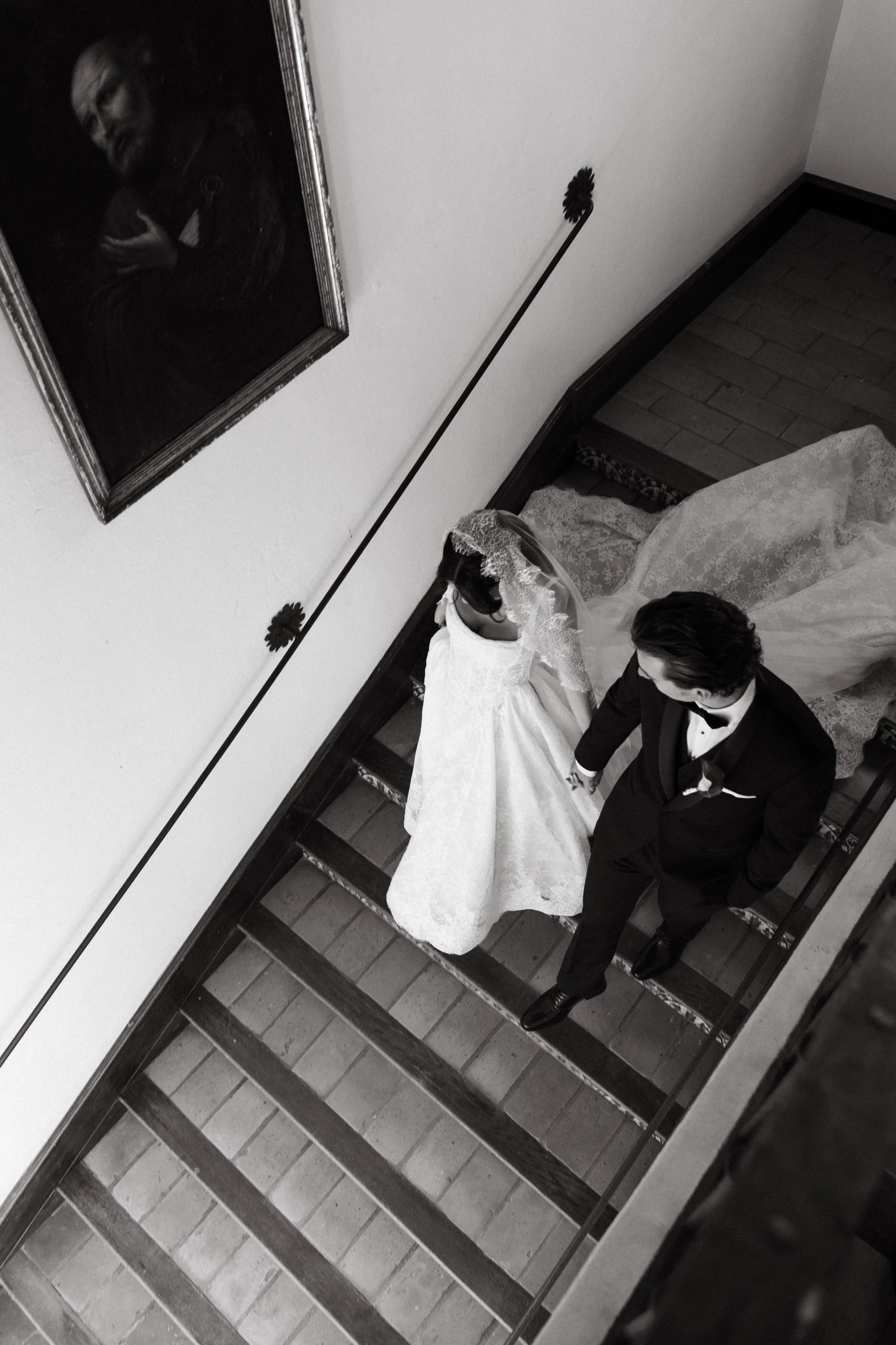 A bride and groom hold hands as they walk down a staircase, with the bride wearing a lace wedding gown and veil, and the groom in a tuxedo. A framed portrait hangs on the wall above them captured in Finca Monasterio Sotogrande Spain