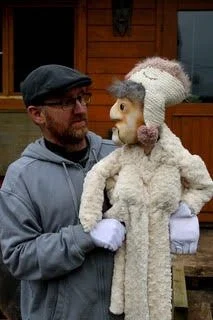 Man holding a puppet resembling an elderly woman dressed in a cozy coat and hat