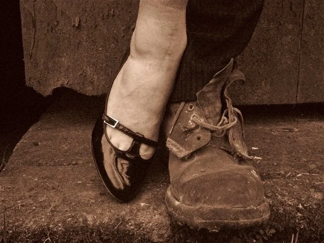 A person wearing a dirty, worn-out work boot on one foot and a clean, shiny, black ballet flat on the other foot, standing on a rough concrete surface.