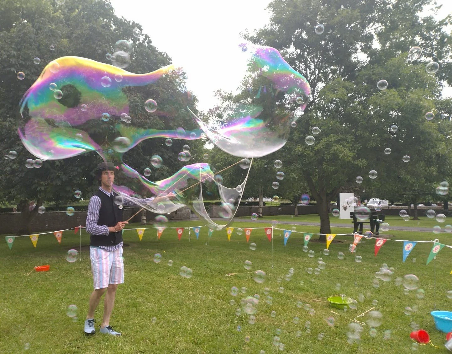 A person holding two large iridescent soap bubbles outside in a park with trees and a grassy area, with colorful pennant banners in the background.