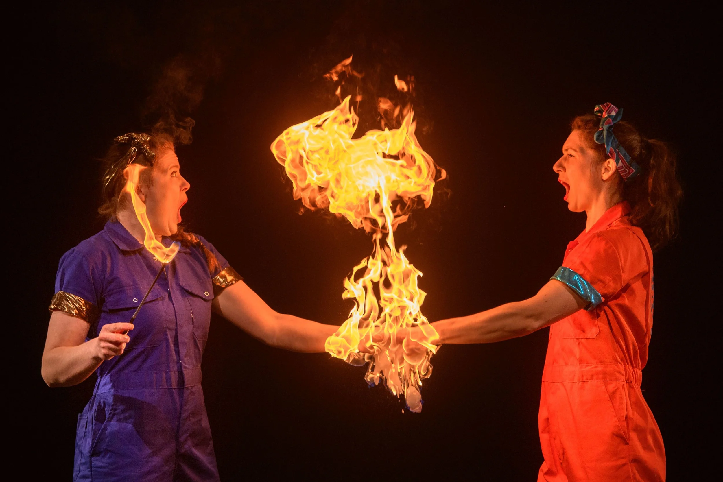 Two women wearing colorful coveralls and headbands are performing a fire stunt at night, with flames connecting their hands.