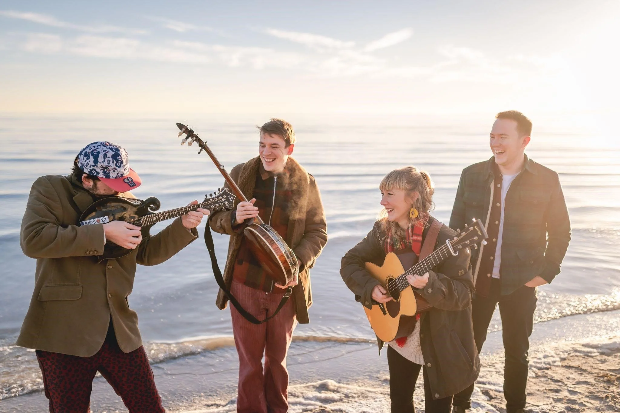 A group of four friends playing musical instruments and singing on a beach during sunset.