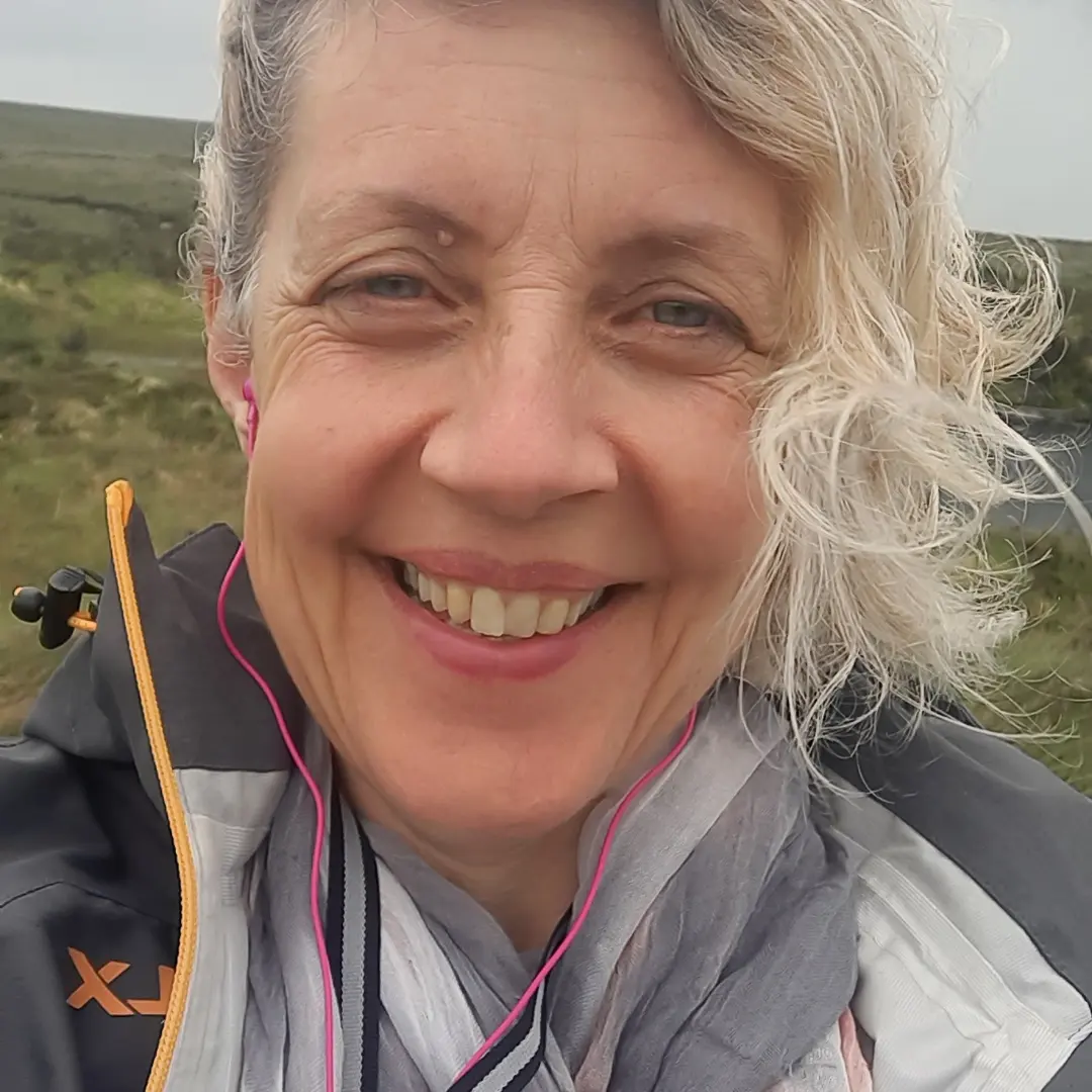 Close-up of smiling woman with curly blonde hair, wearing a jacket and pink earphones outdoors in a grassy landscape.