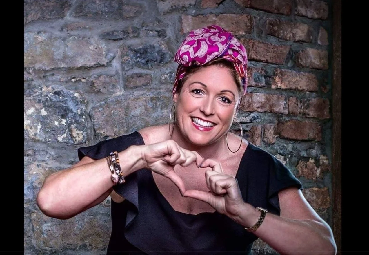 A woman with a pink patterned headscarf and hoop earrings smiling and making a heart shape with her hands in front of a brick wall.