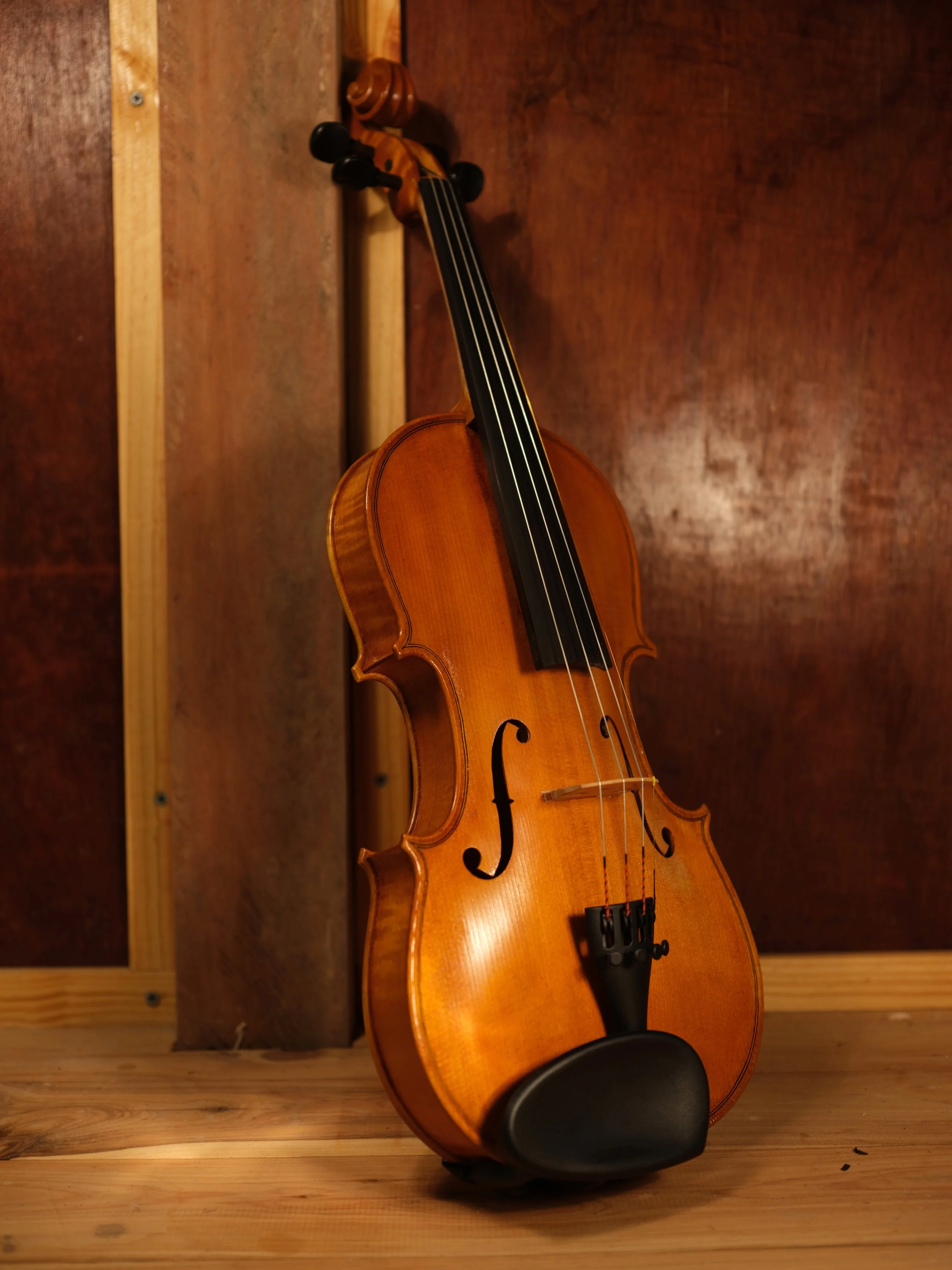 A violin with a wooden body and black fingerboard resting against a wood-paneled wall on a wooden floor.