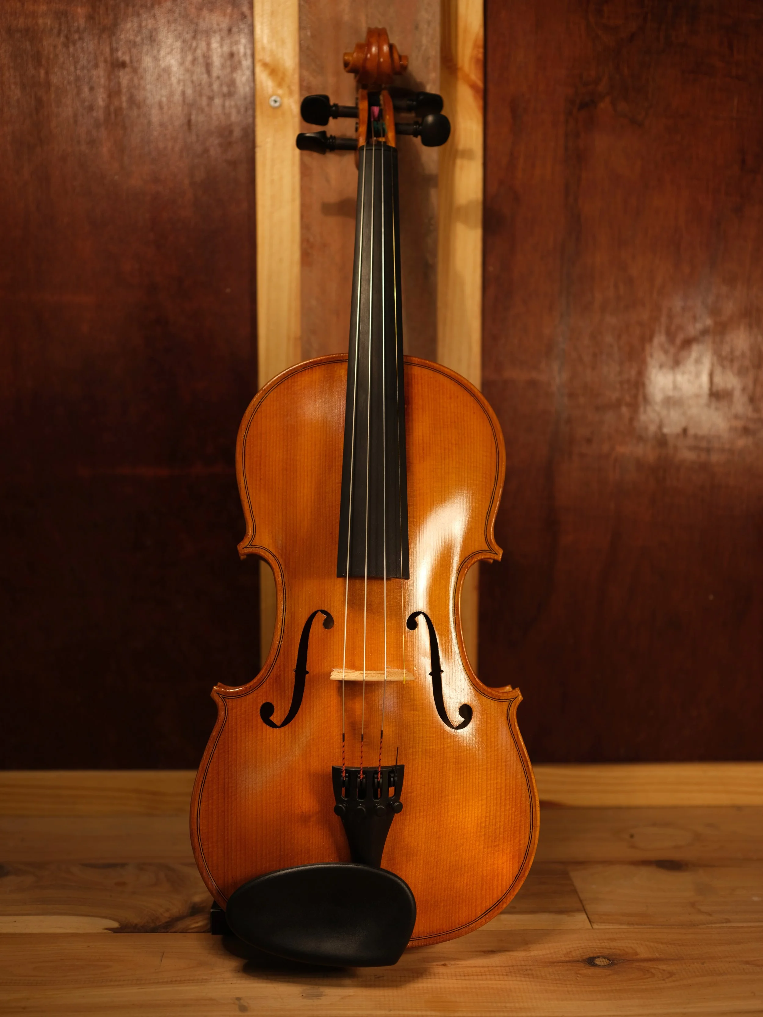 A wooden violin with a black chin rest and four strings, standing upright against a wooden wall.