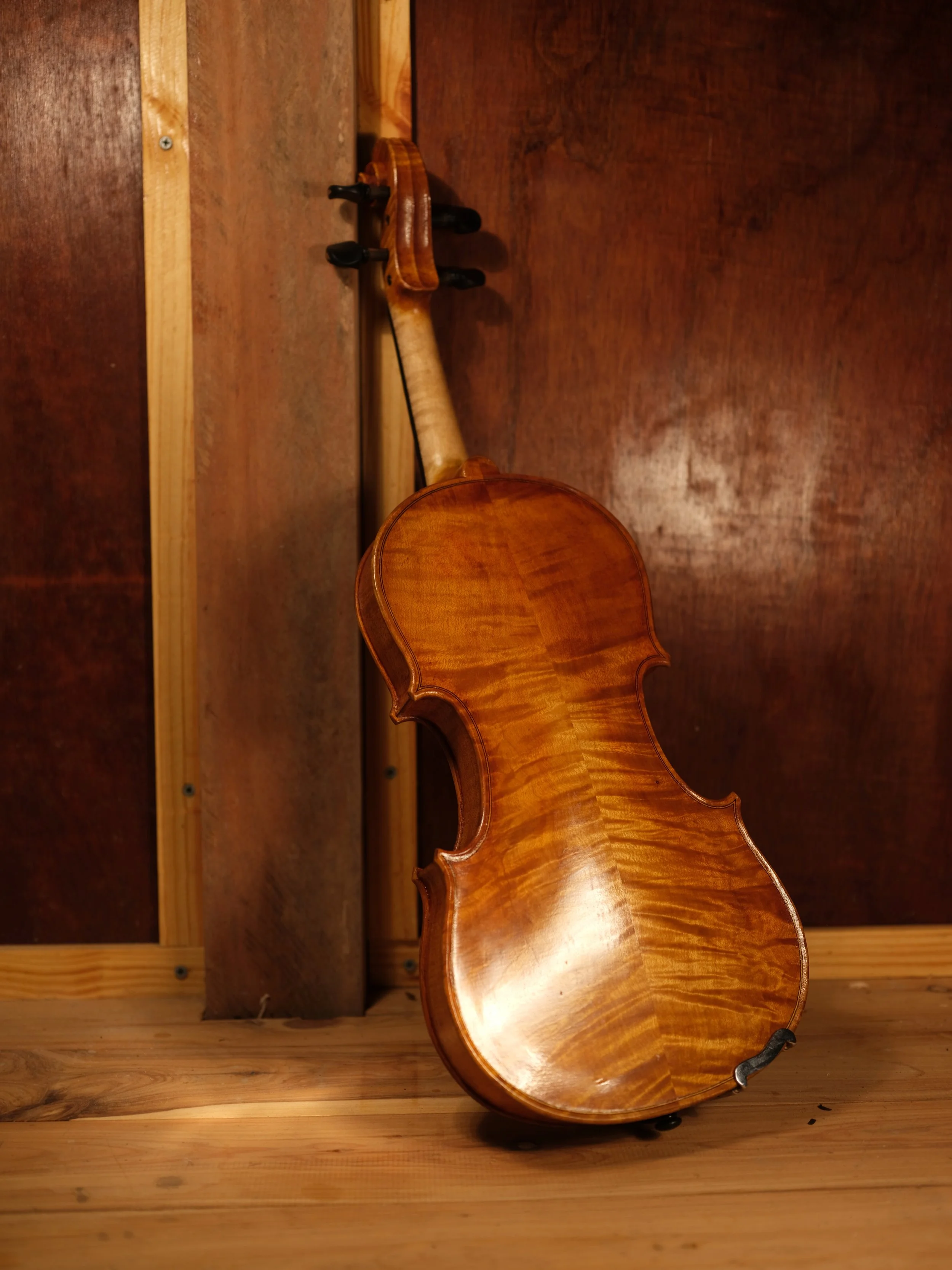 A polished wooden violin leaning against a wall on a wooden floor.