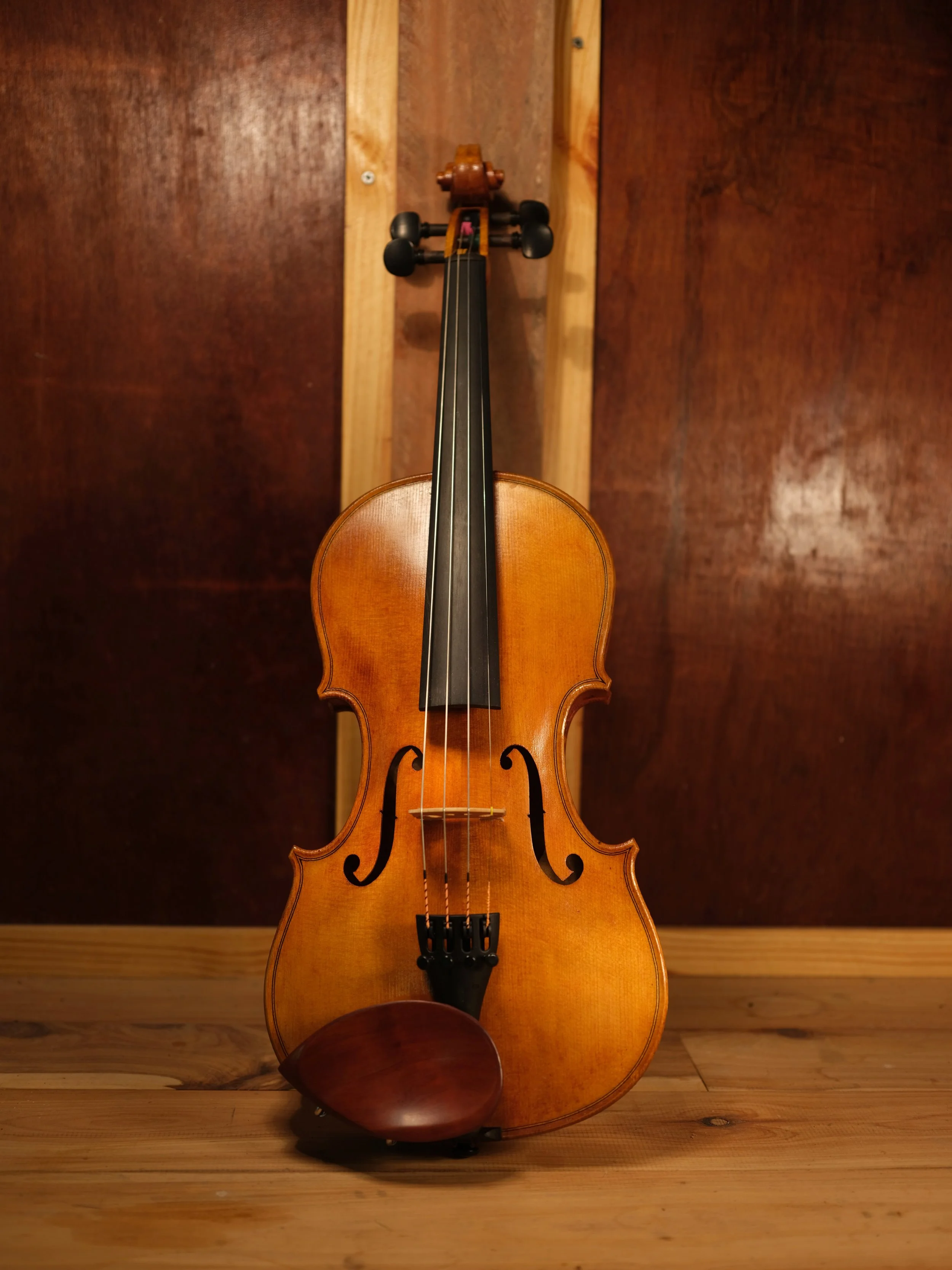 A wooden violin with a dark brown chin rest, three strings, and black tuning pegs, standing upright against a wooden wall.
