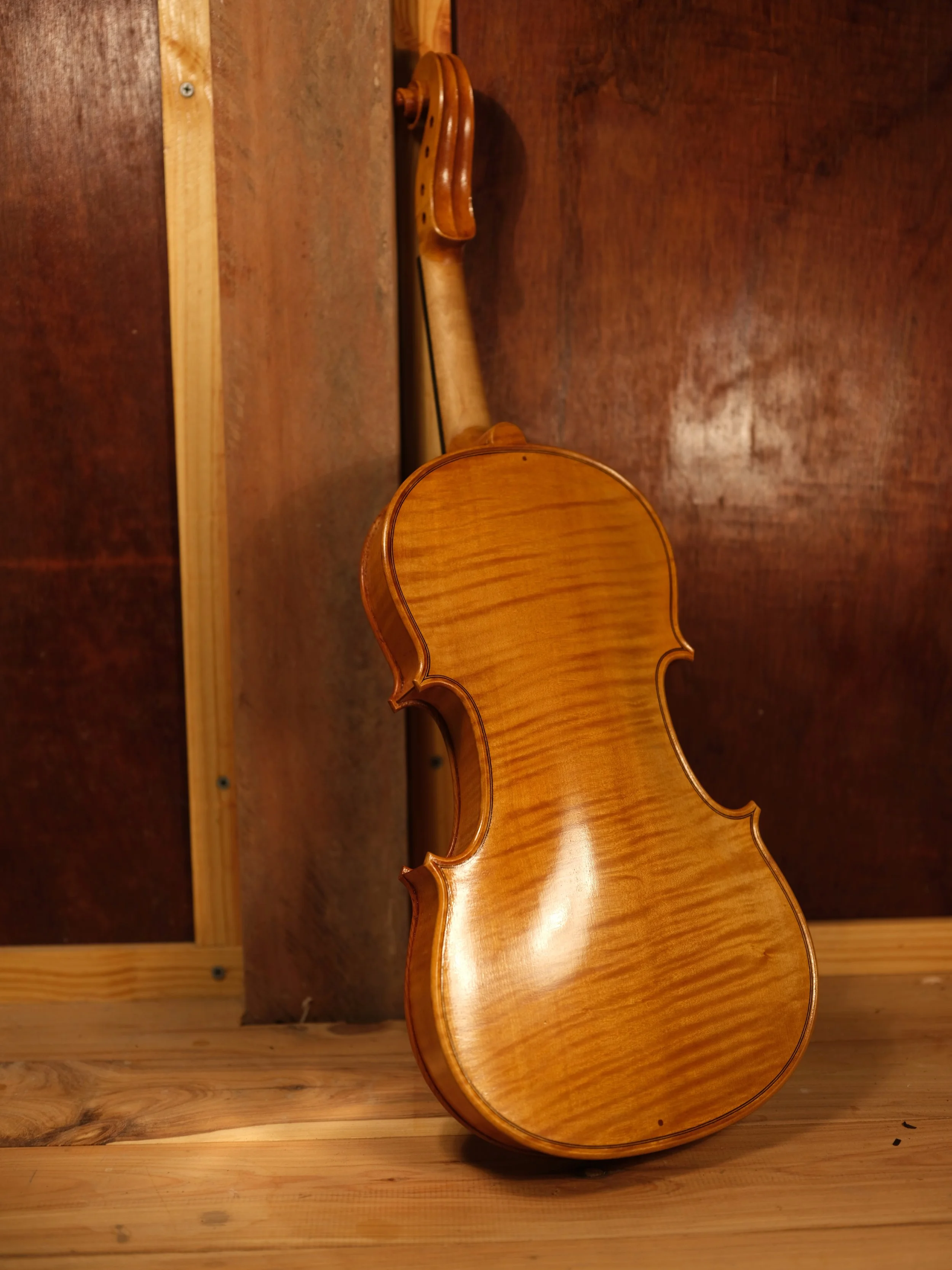 A wooden violin leaning against a wooden wall on a wooden floor.