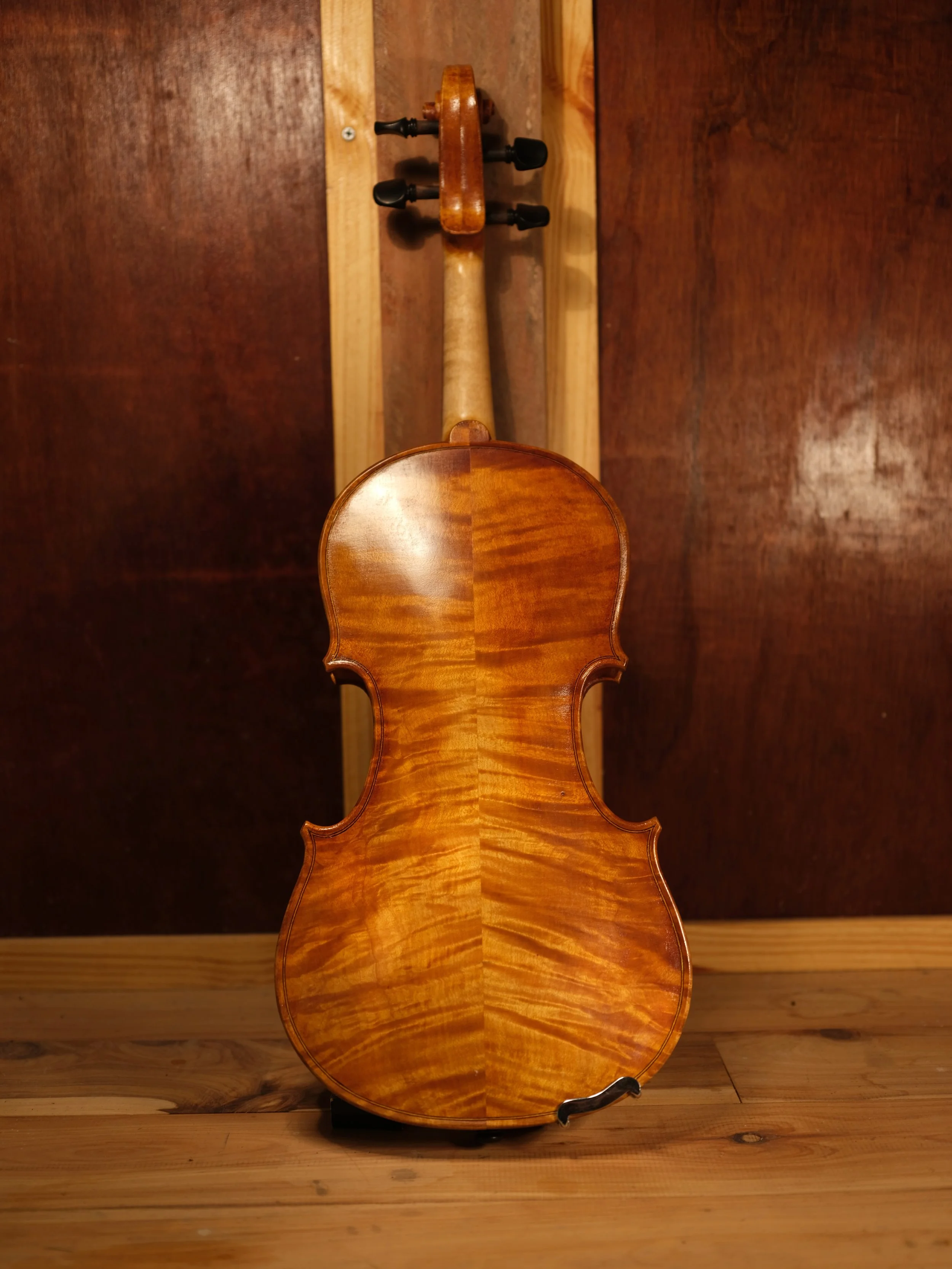 The image shows a wooden violin placed with its back facing up, standing on a wooden floor against a wooden wall. The violin's back has a highly polished, natural wood finish with visible wood grain patterns.