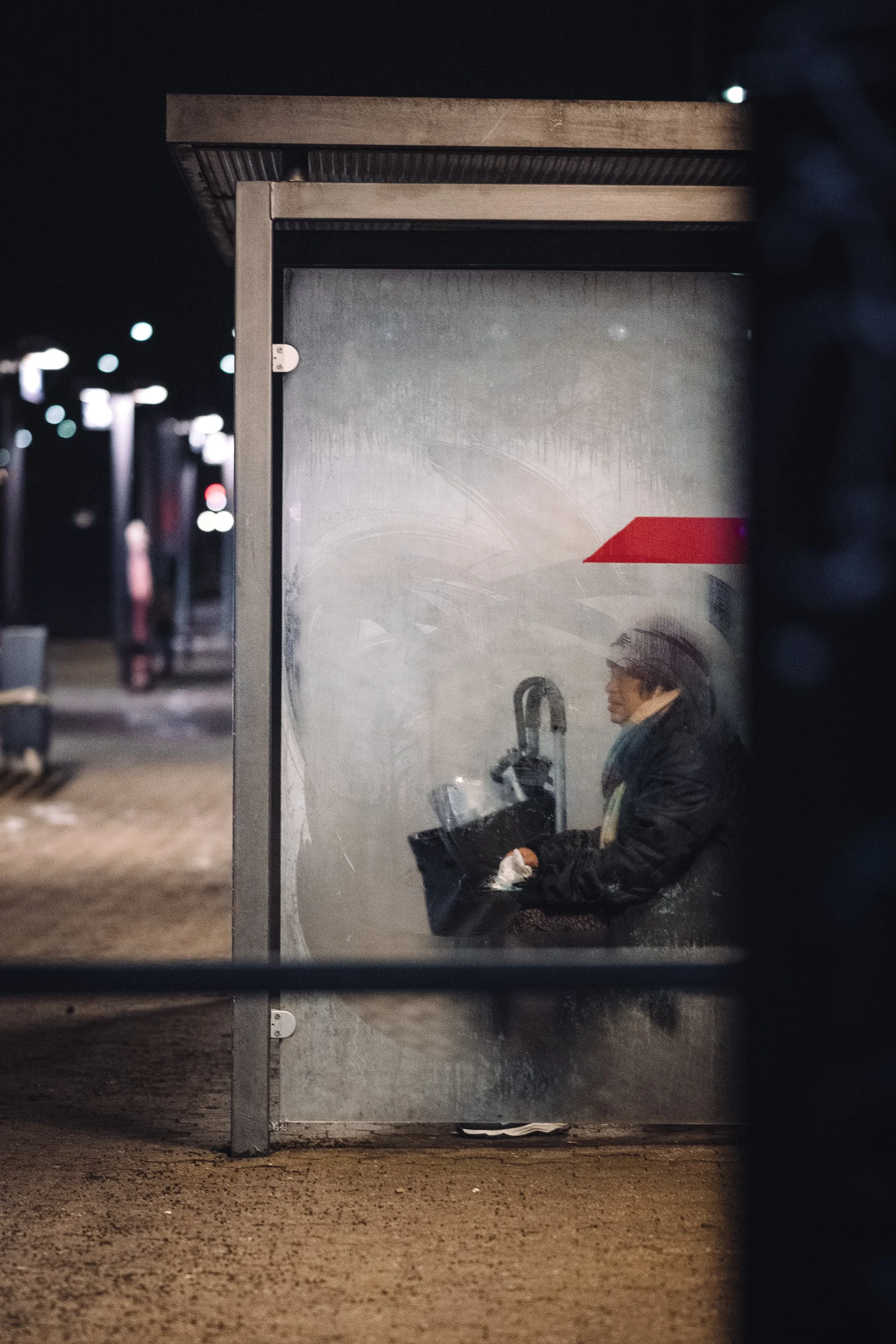 A person in a dark jacket and glasses sitting alone at a bus stop at night, reading a book with a bag on their lap, surrounded by a dimly lit urban environment.