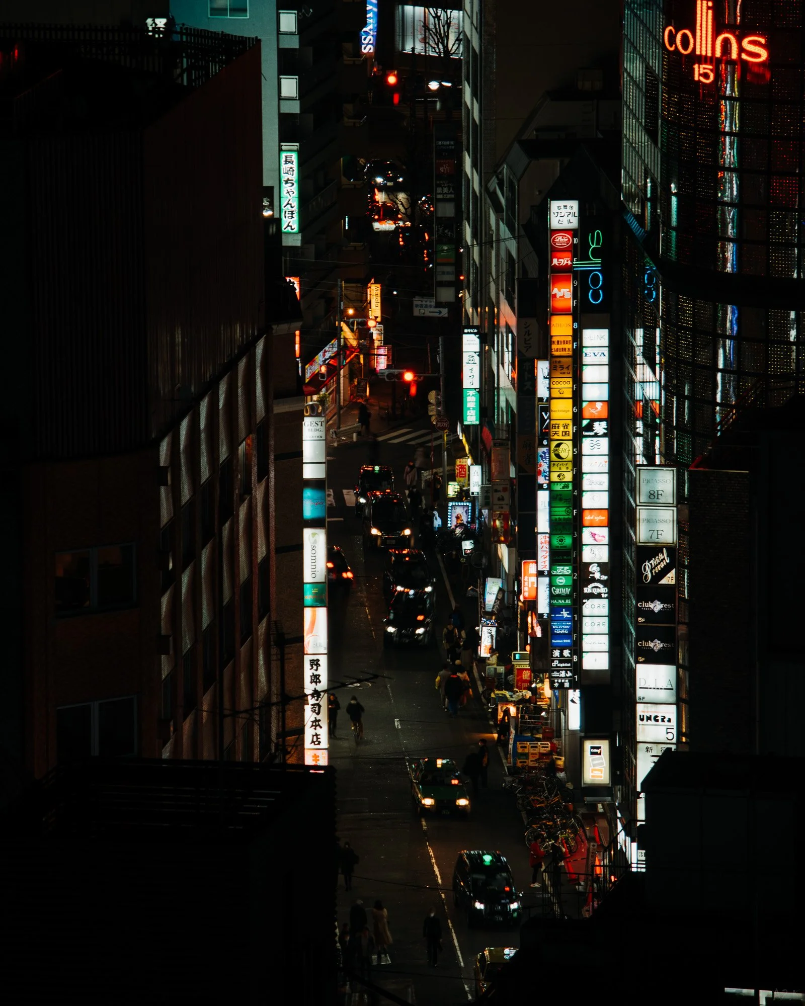 Nighttime city street with neon signs and illuminated buildings, cars, and pedestrians.