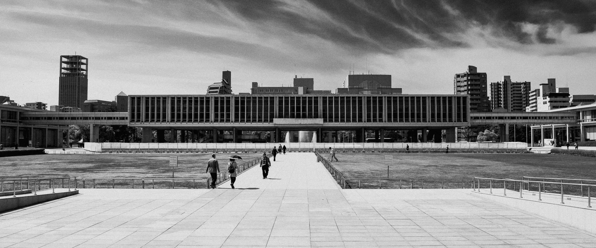 Black and white photo of a cityscape with tall buildings in the background and people walking on a wide walkway in the foreground.