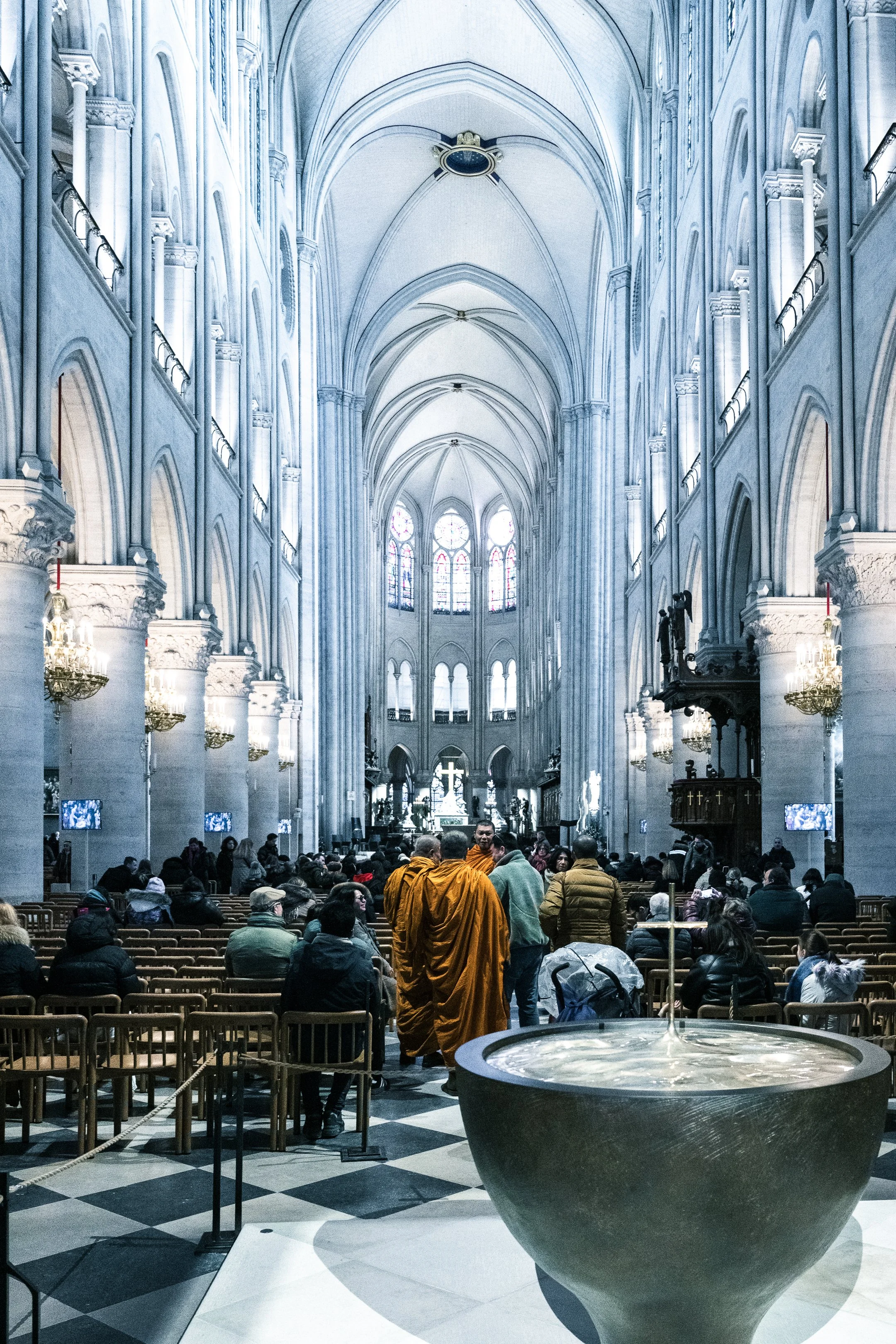 Interior view of the high vaulted ceilings, stained glass windows, and ornate chandeliers of Notre Dame De Paris. People are seated and standing inside, with buddist monks in orange robes at the center.
