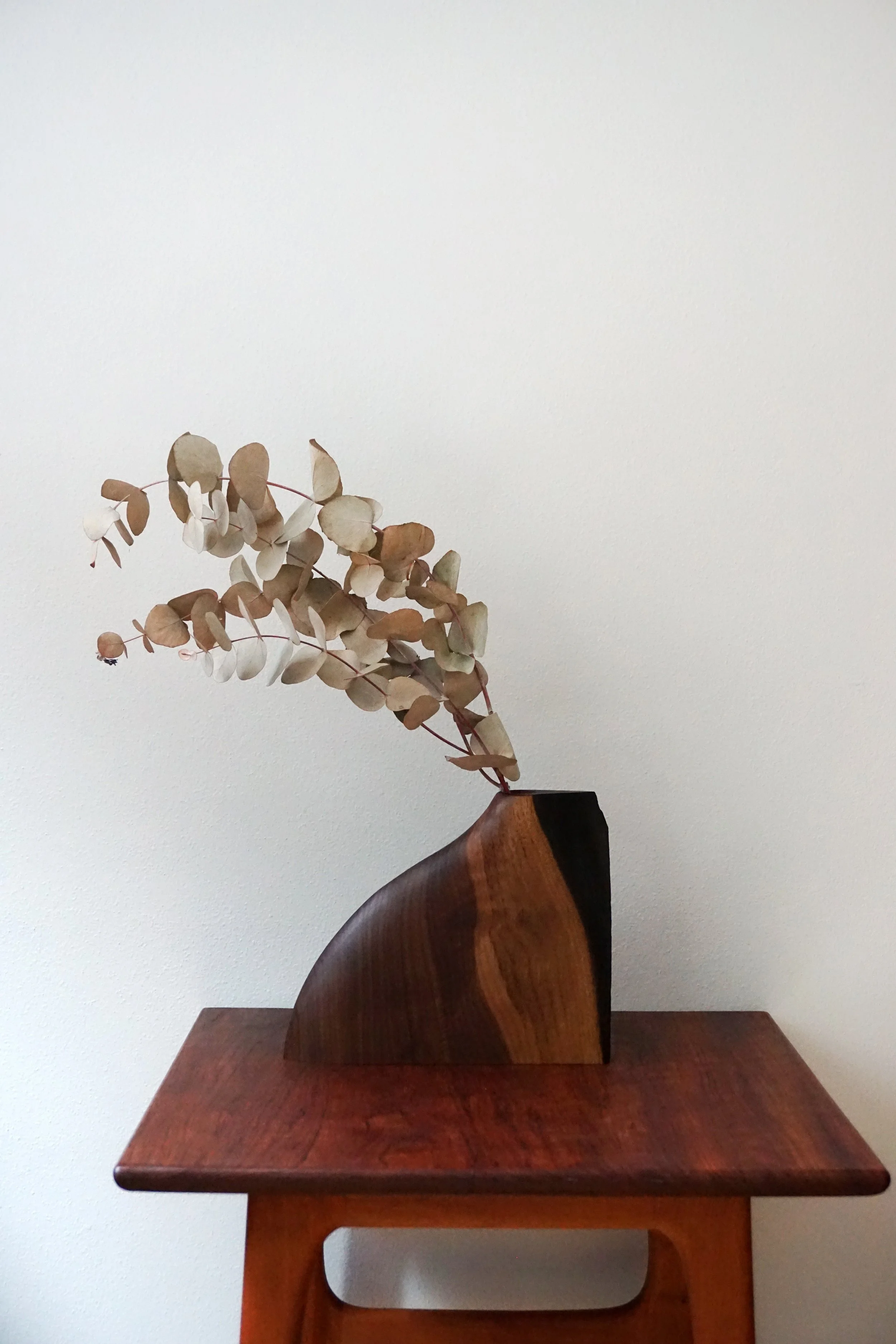 A wooden sculpture of a vase with dried leaves or flowers on top, placed on a wooden table against a white wall.