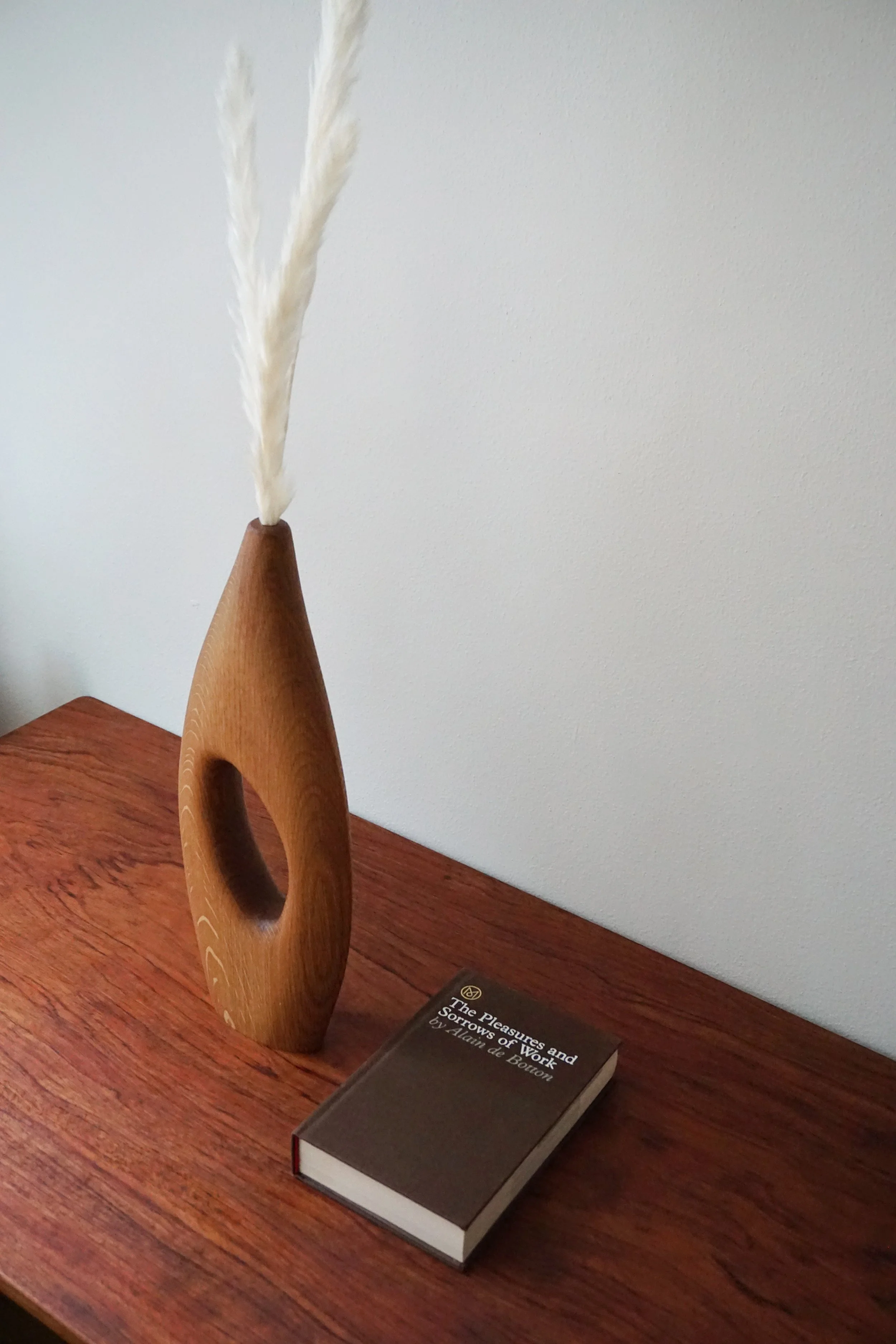 A wooden sculpture with an elongated oval opening, holding white pampas grass. Next to the sculpture is a closed book titled "The Pleasures and Sorrows of Work" by Alain de Botton, placed on a wooden table.