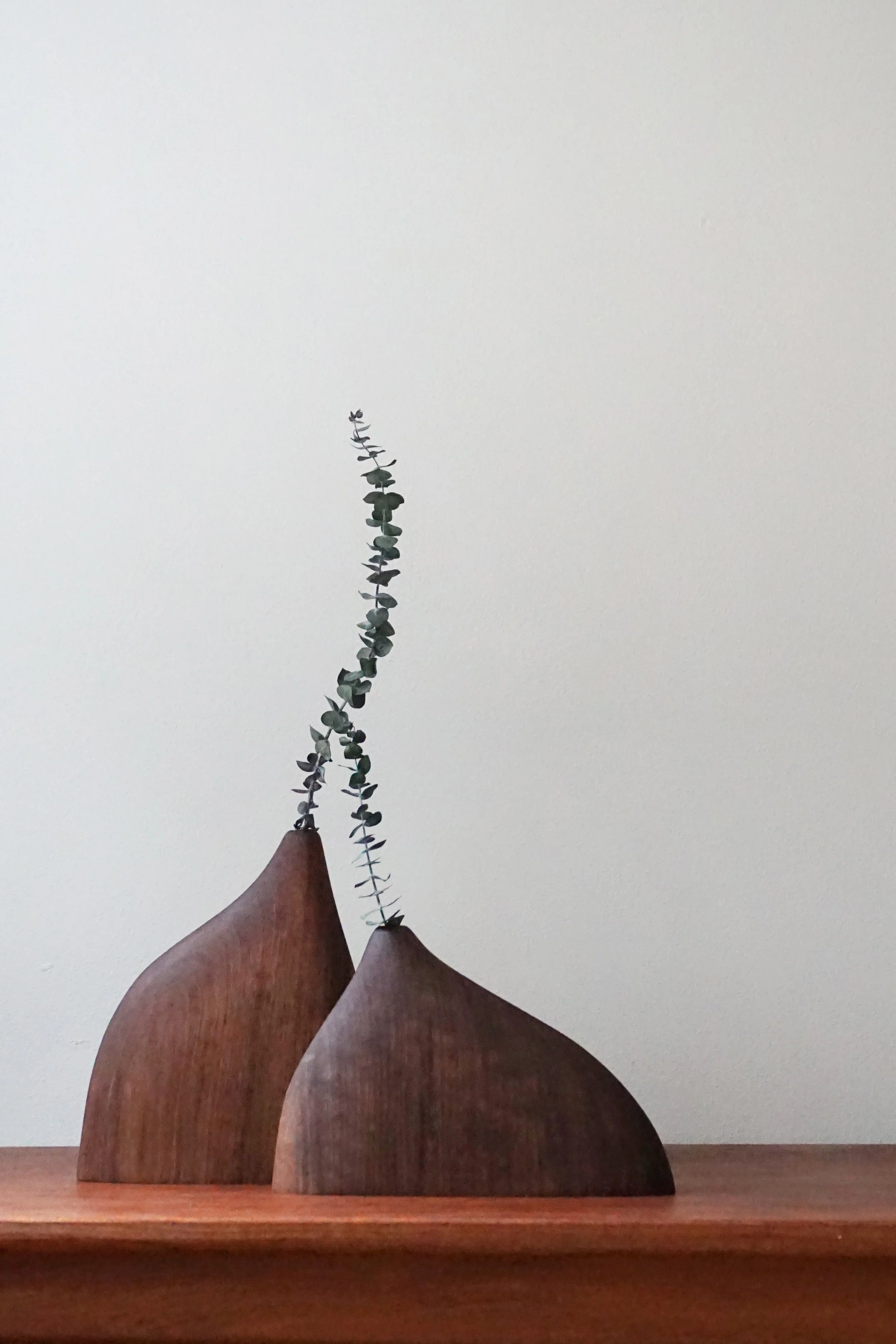 Three wooden bird-shaped sculptures with a eucalyptus branch on top, placed on a wooden surface against a plain wall.