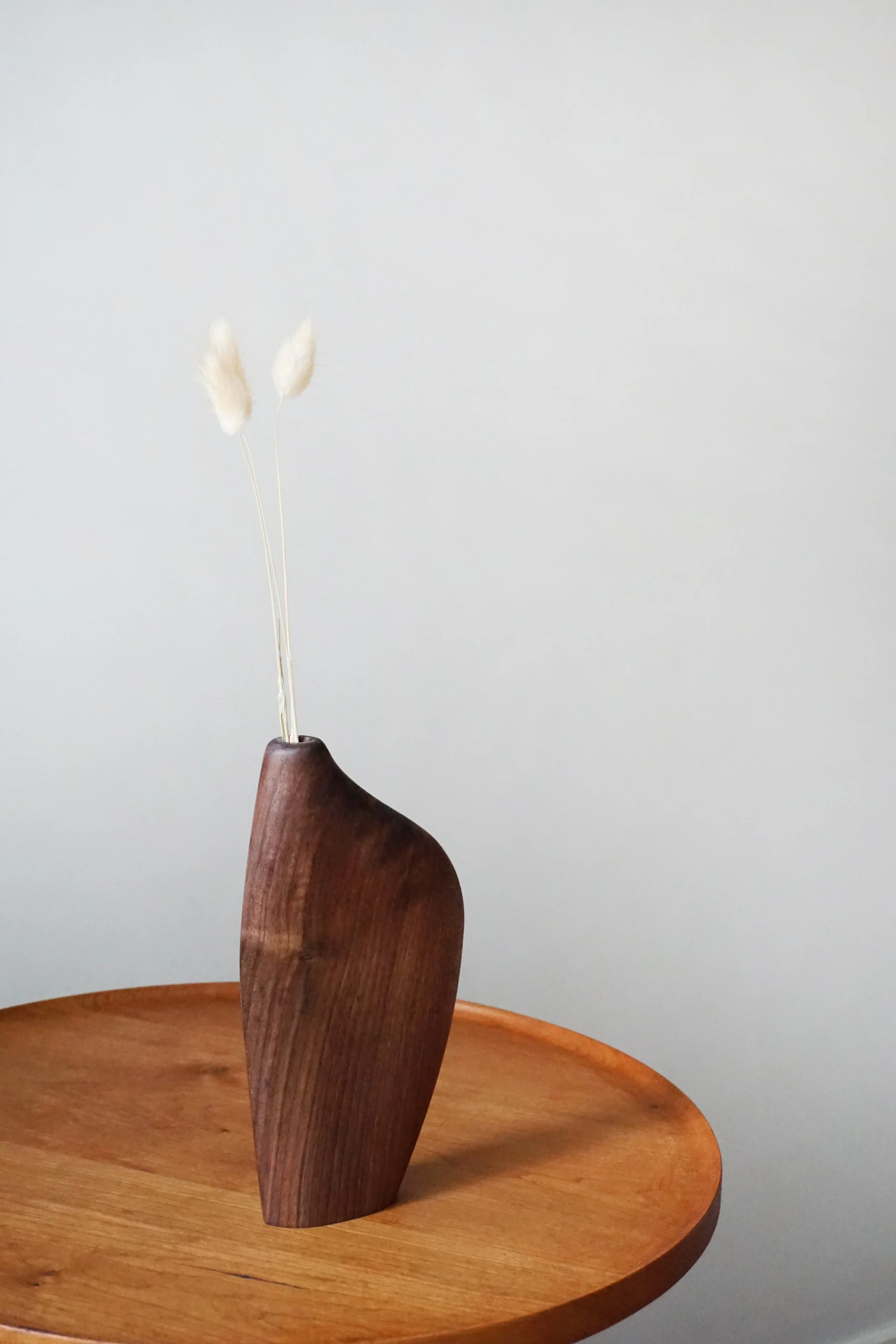A wooden bowl with a wooden stand holding a dried plant with two fluffy white seed heads.