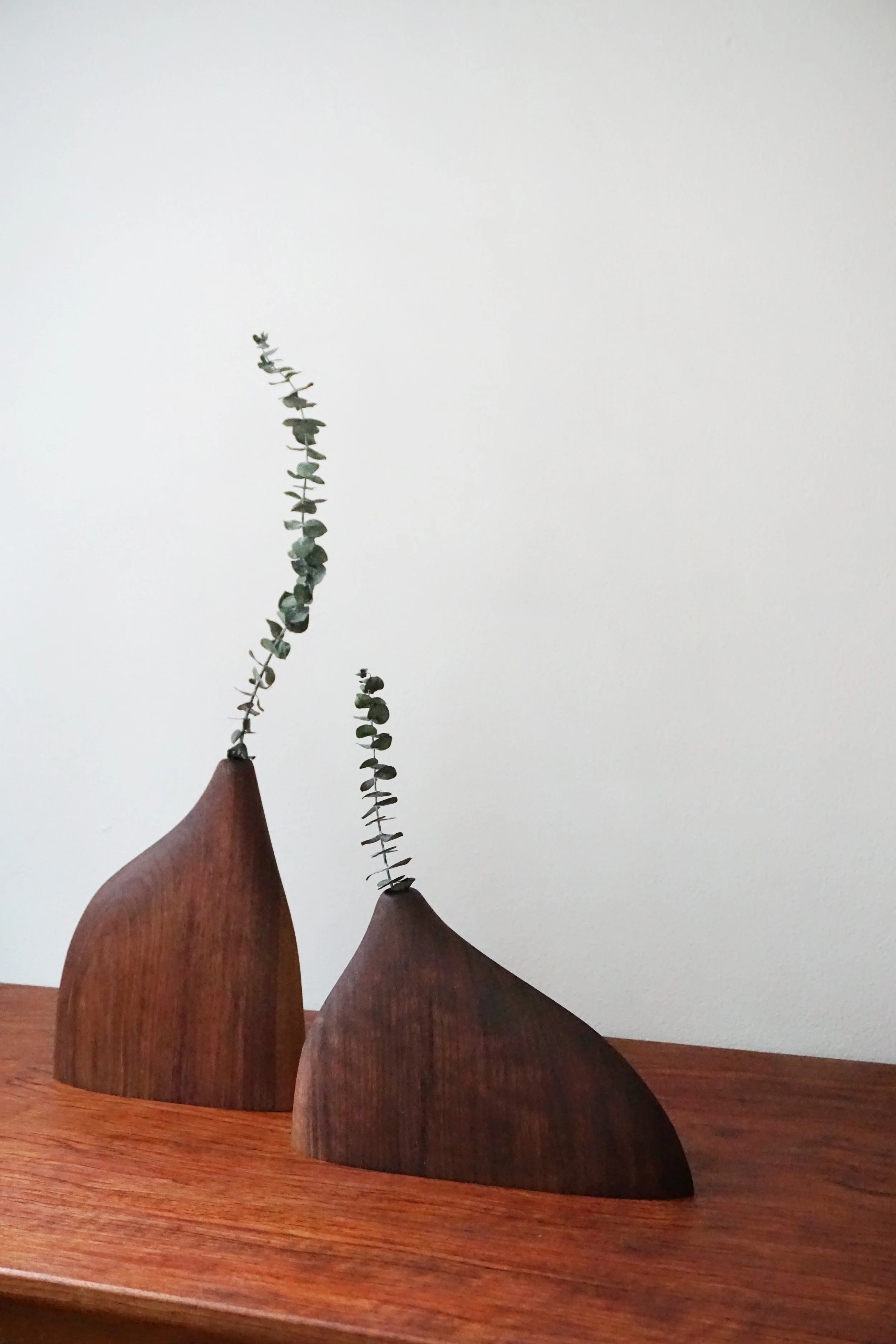 Three wooden sculptures with rounded top shapes placed on a wooden surface, holding tall, thin branches with small green leaves against a plain white wall background.