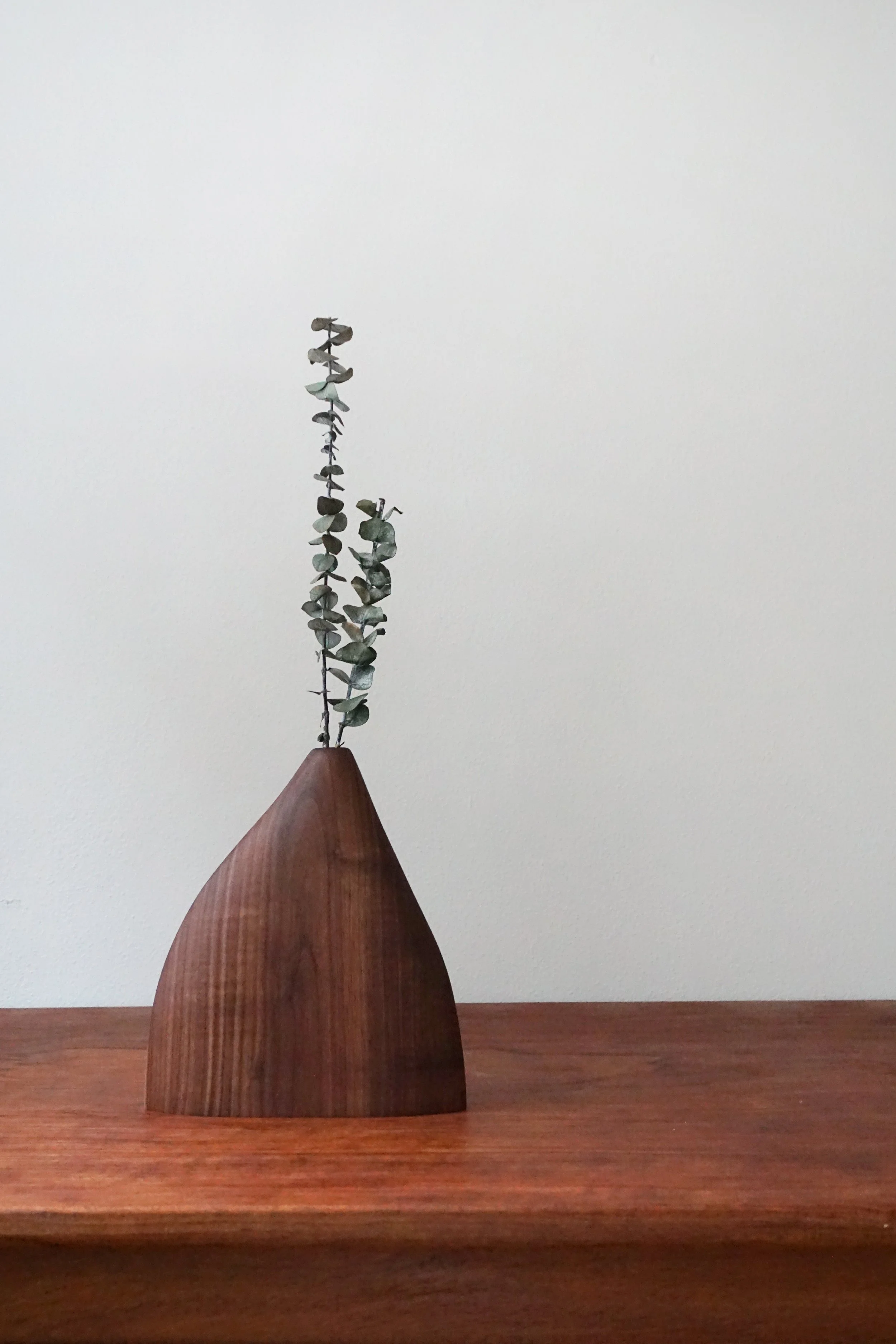 A wooden sculpture resembling a cone with a tall, thin, metal sculpture of a branch with leaves on top, placed on a wooden surface with a plain white wall in the background.