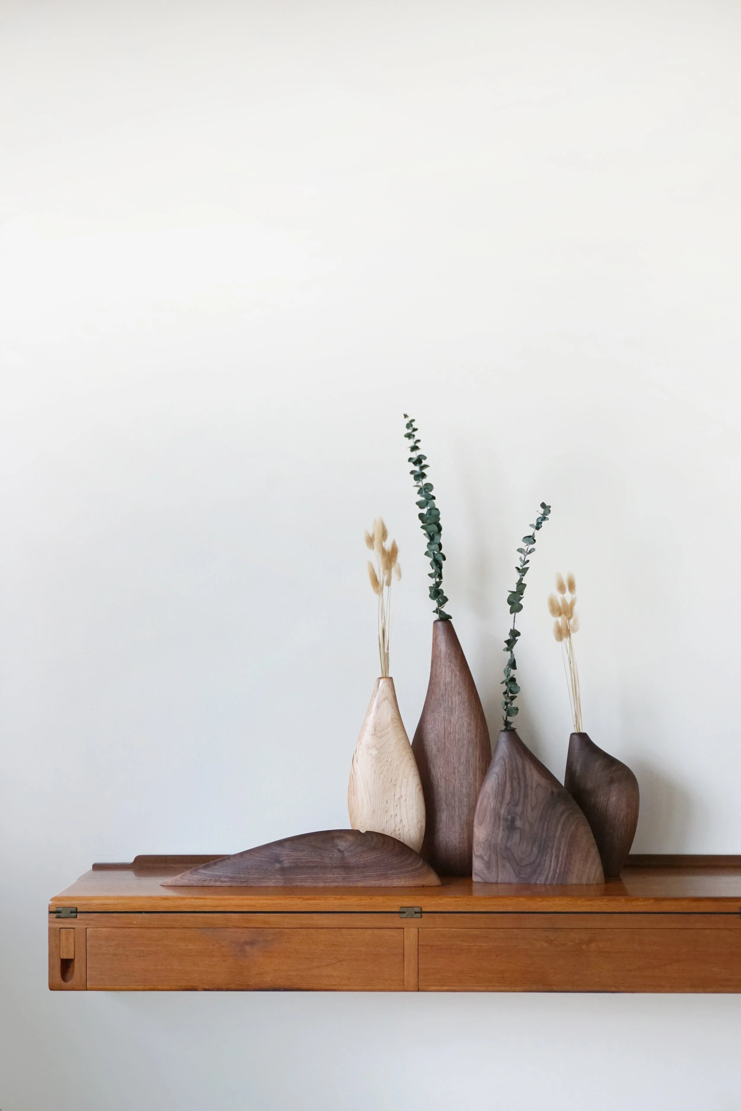 Wooden vases with dried and green foliage on a floating wooden shelf.