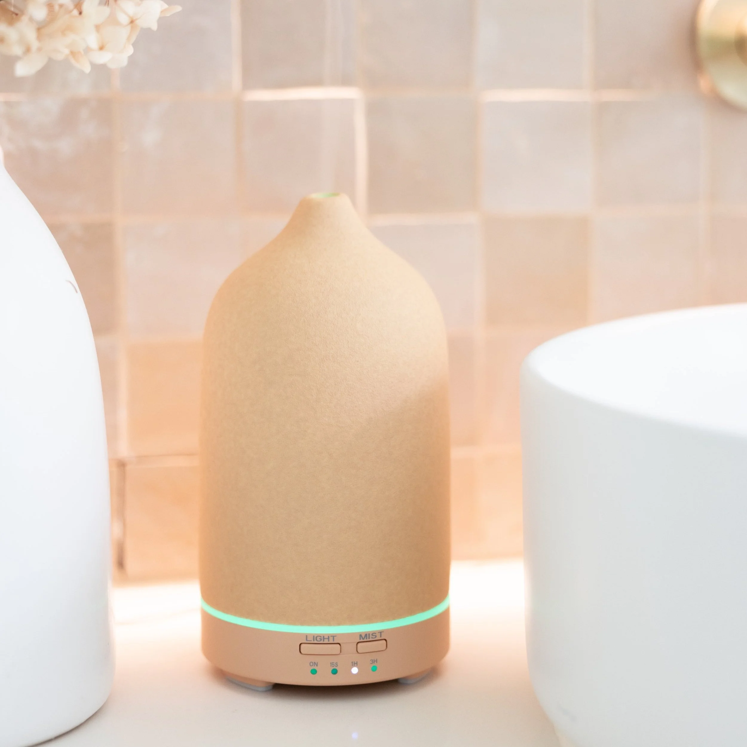 A tan essential oil diffuser with a modern design, featuring buttons labeled 'Light' and 'Mist', sits on a bathroom counter next to a white vase and a tiled backsplash.
