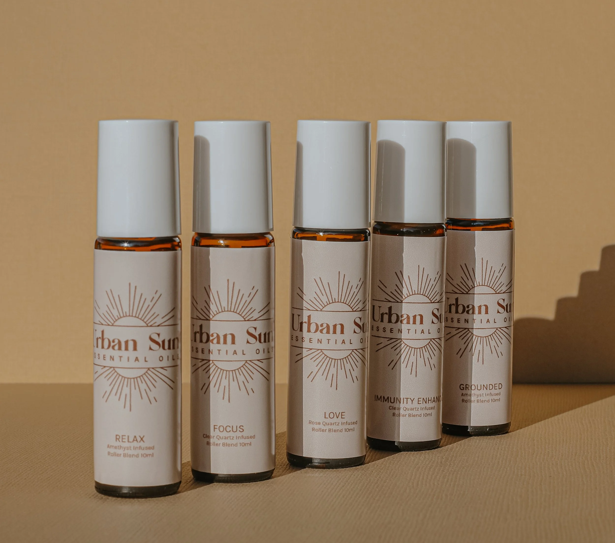 Five amber glass roller bottles of Urban Sun Essential Oils labeled Relax, Focus, Love, Immunity Enhanced, and Grounded against a beige background.