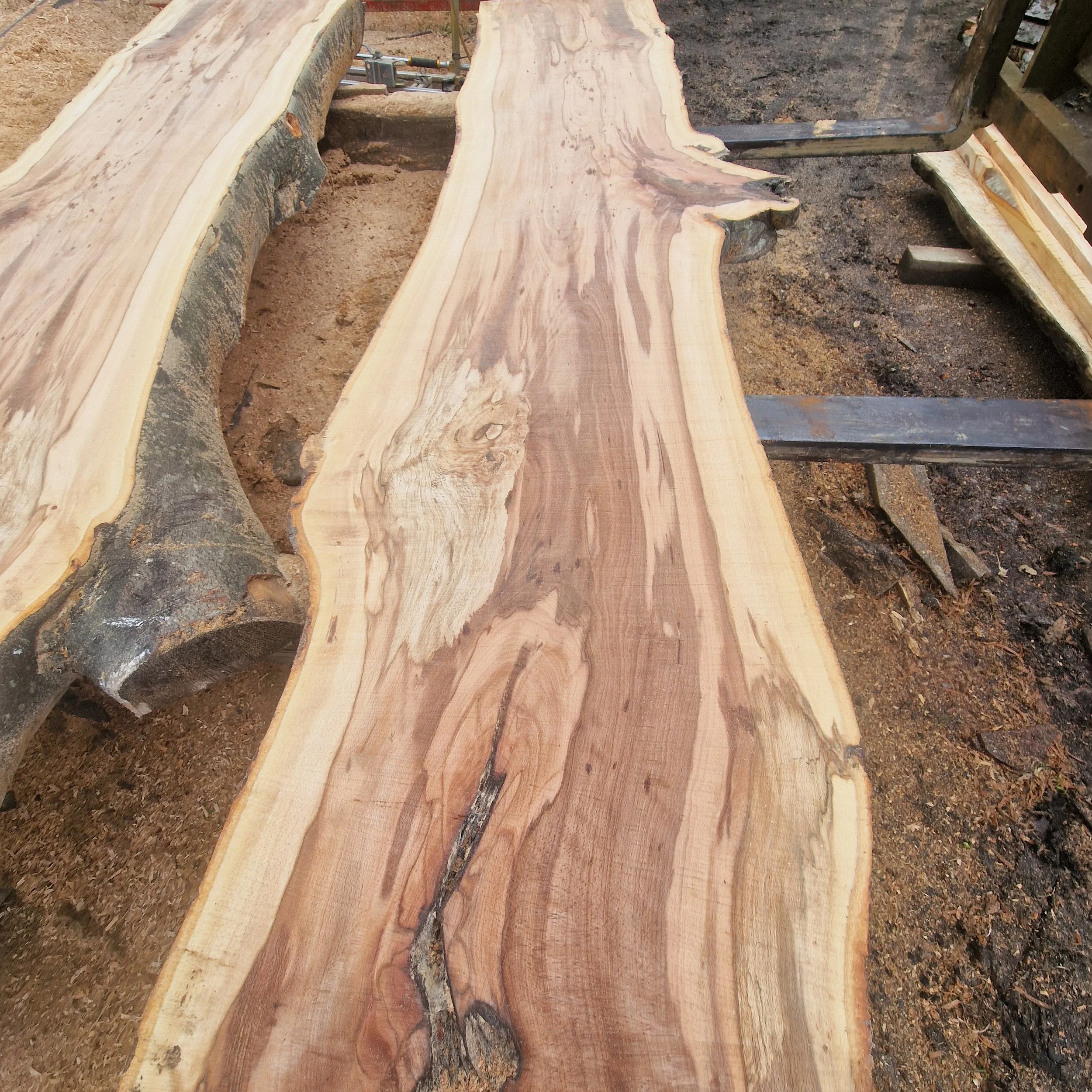 A long, live-edge wooden slab with a natural finish, placed on a construction site or workshop, with visible grain and irregular edges.