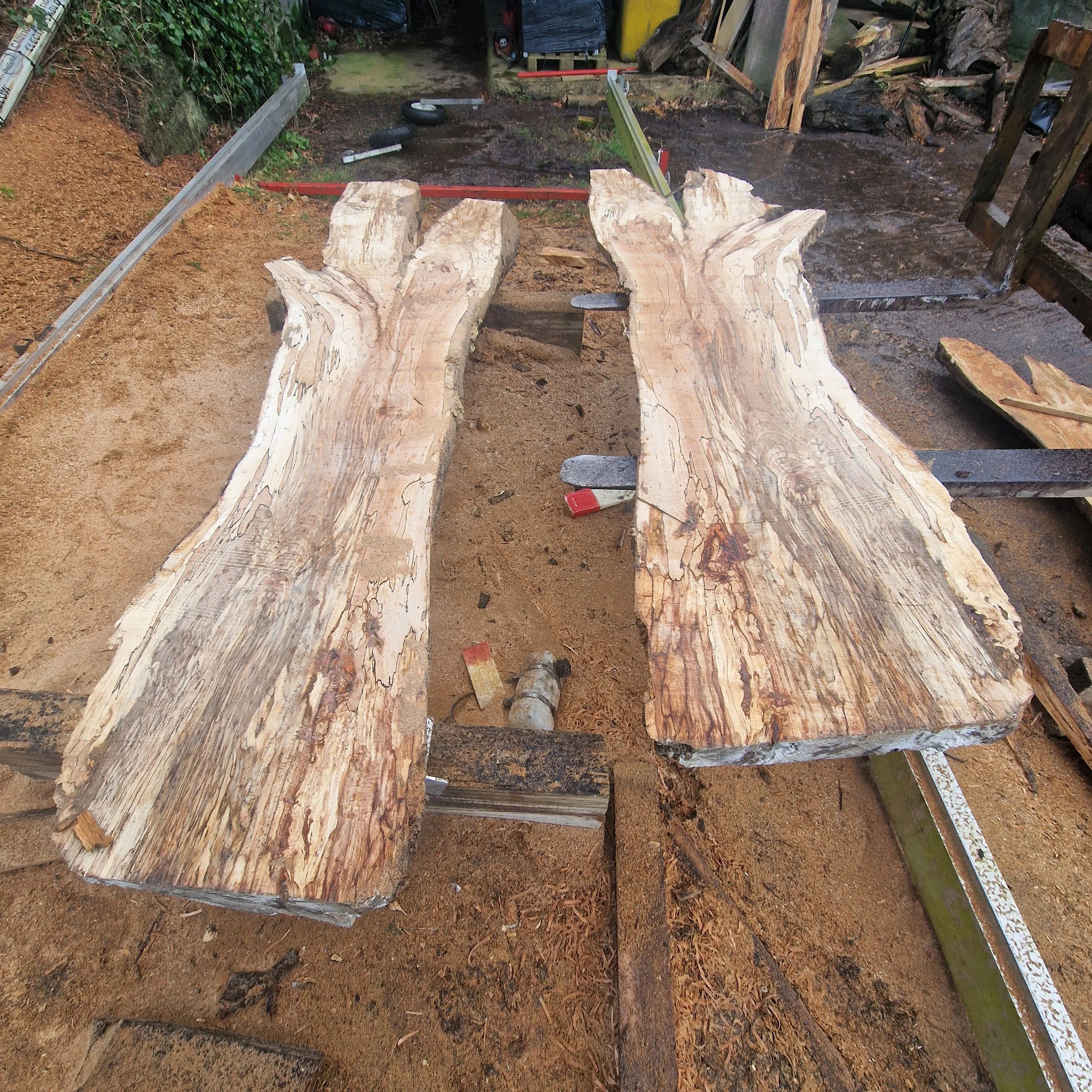 Two large, roughly carved wooden slabs are placed on a sawhorse in a woodworking shed or outdoor workshop, with sand and various tools around.